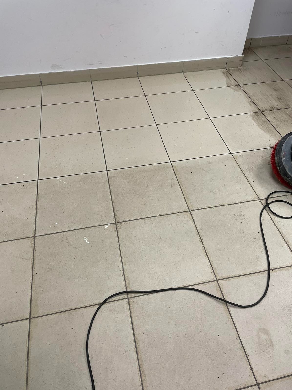 Maszyna do czyszczenia posadzek w narożniku pomieszczenia, widoczne ślady czyszczenia na kafelkach podłogowych.