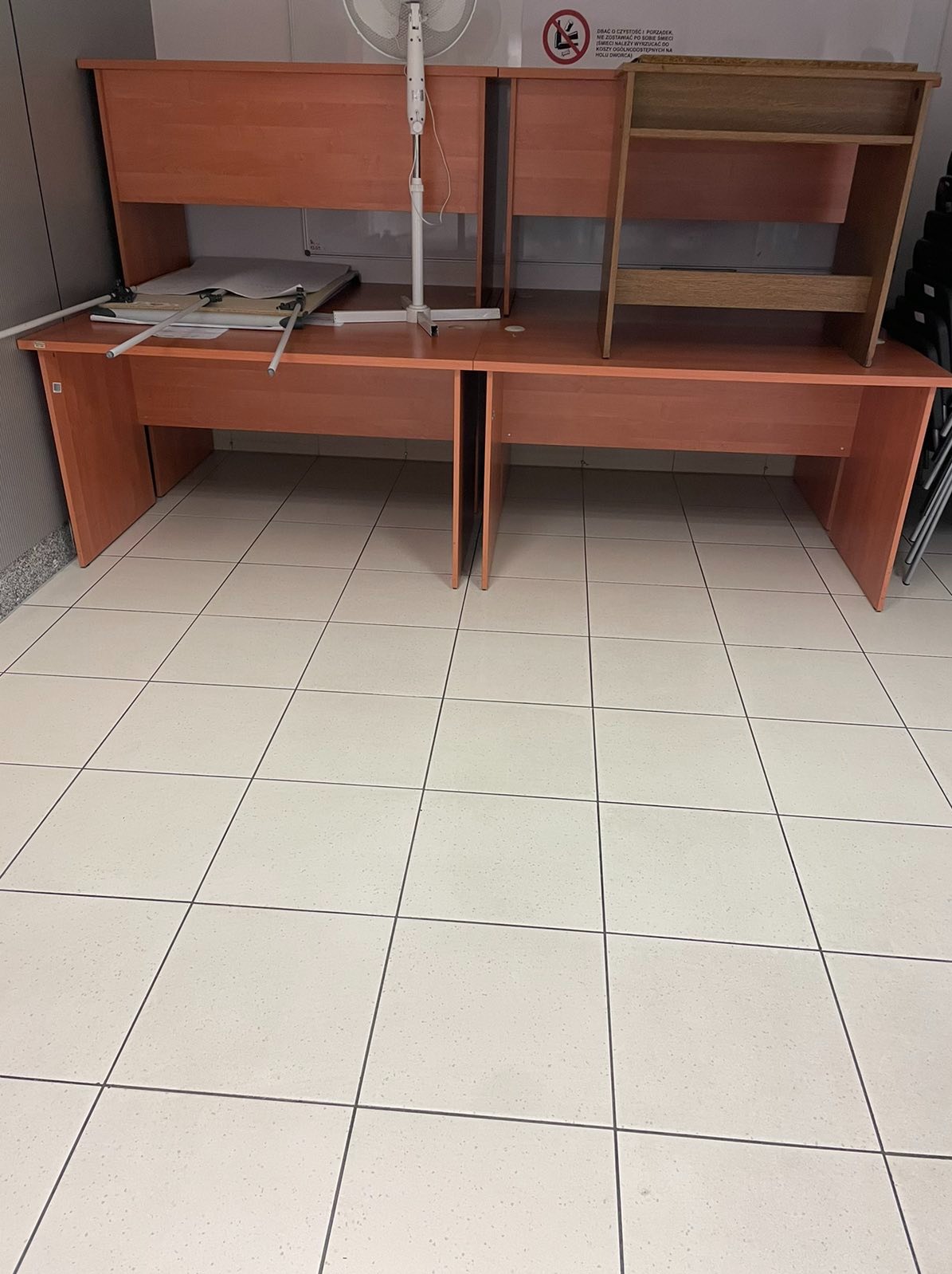 Dwa drewniane biurka ustawione obok siebie na kafelkach, z widocznym stojącym wentylatorem i półkami na jednym z biurek. Na jednym z biurek leżą rozłożone papiery i metalowe elementy.