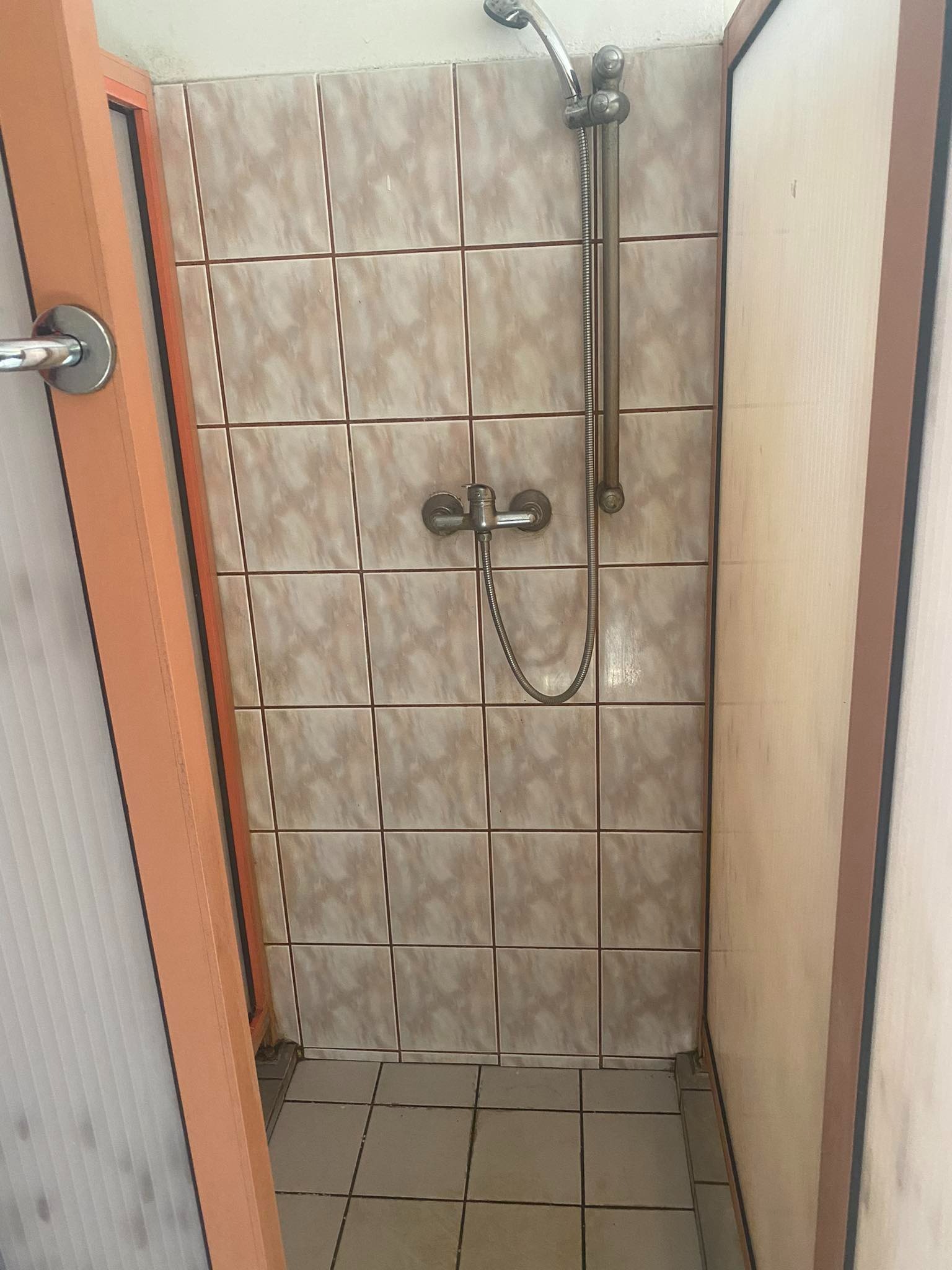 Wnętrze kabiny prysznicowej z beżowymi płytkami, widoczna bateria i słuchawka prysznicowa, fragment drzwi z pomarańczową framugą po lewej stronie.