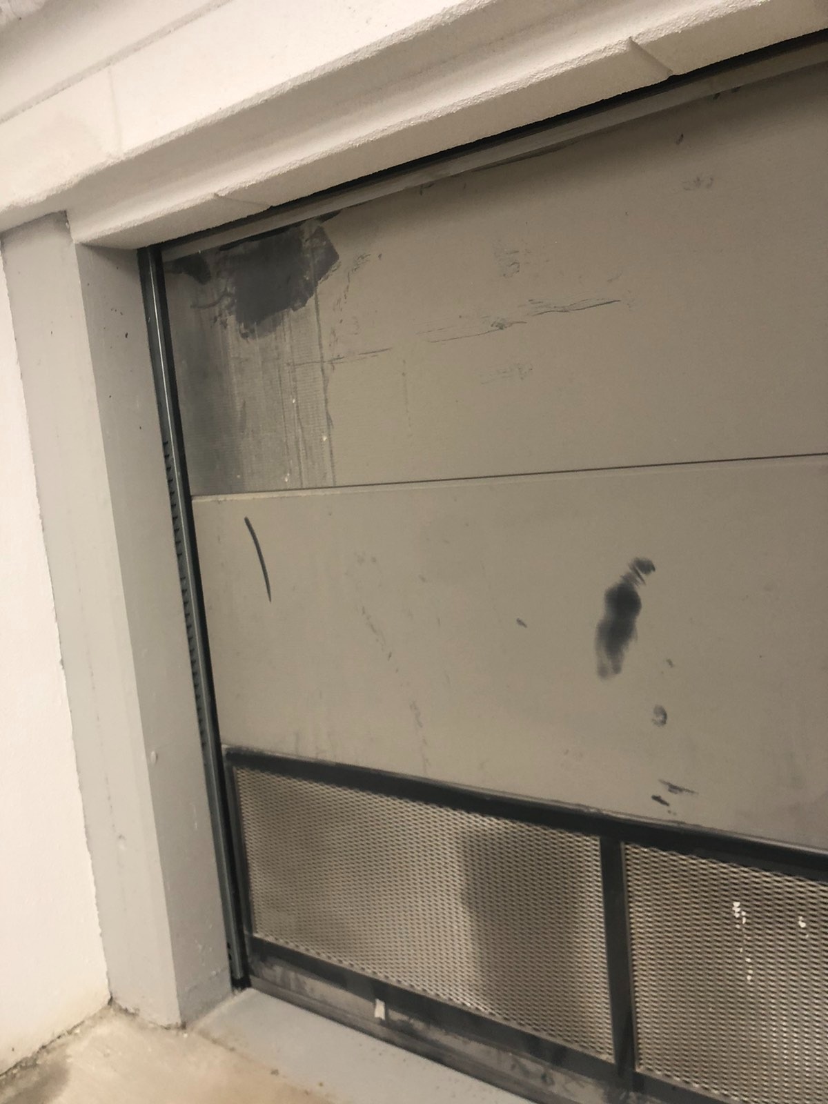 Brudna, szara brama garażowa z metalową siatką w dolnej części, widoczne ślady po zabrudzeniach i zacieki.