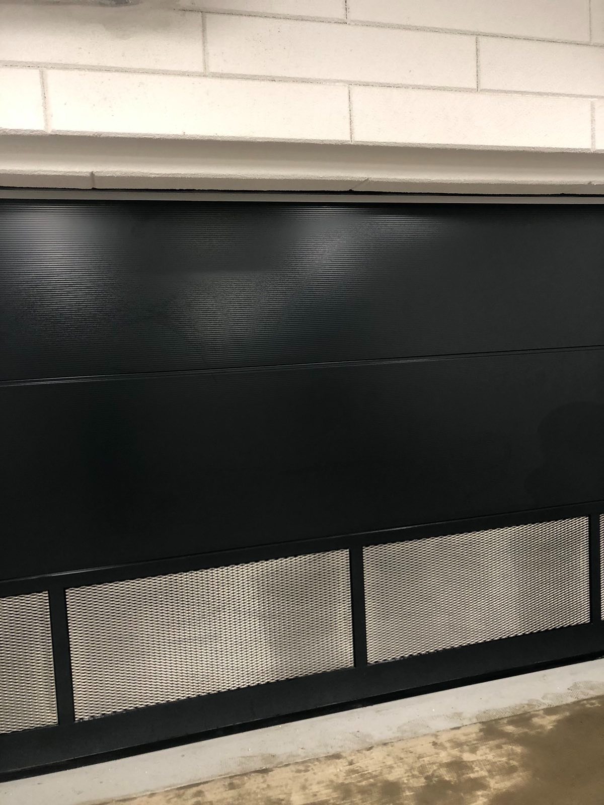 Zamknięta, nowoczesna, czarna brama garażowa segmentowa z perforowanym panelem dolnym, widoczna od wewnątrz, na tle białej ściany z cegieł.