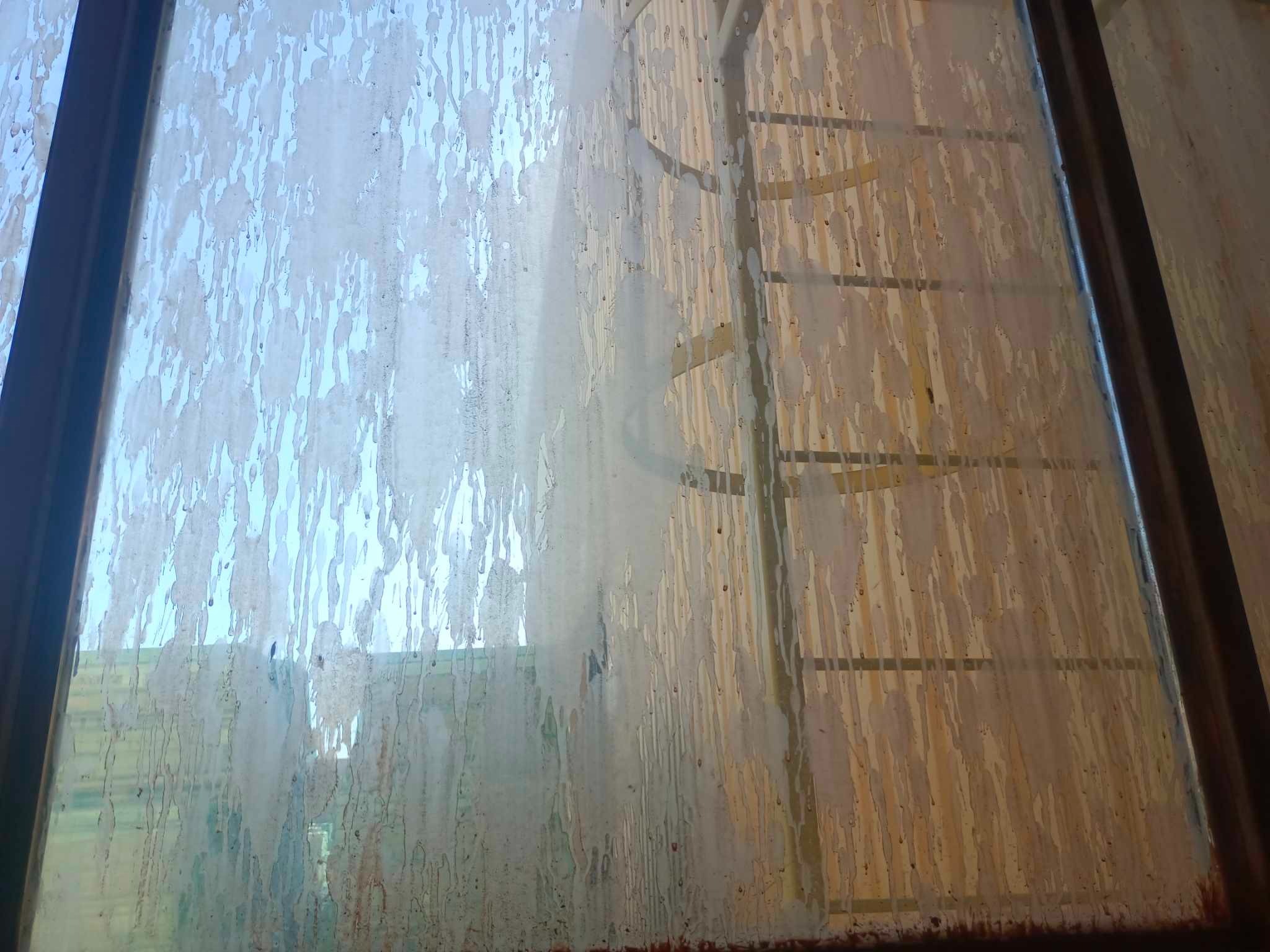 Silnie zabrudzone okno z widocznymi smugami i zaciekami, przez które słabo przebija światło dzienne, z widoczną ramą okienną i fragmentem elewacji budynku za oknem.