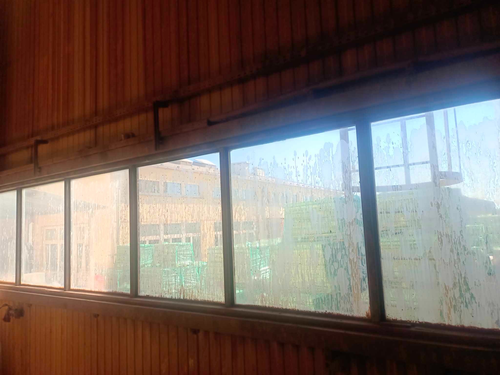 Brudne okna w hali produkcyjnej z drewnianą boazerią, widoczne zacieki i kurz, na zewnątrz budynek i palety.