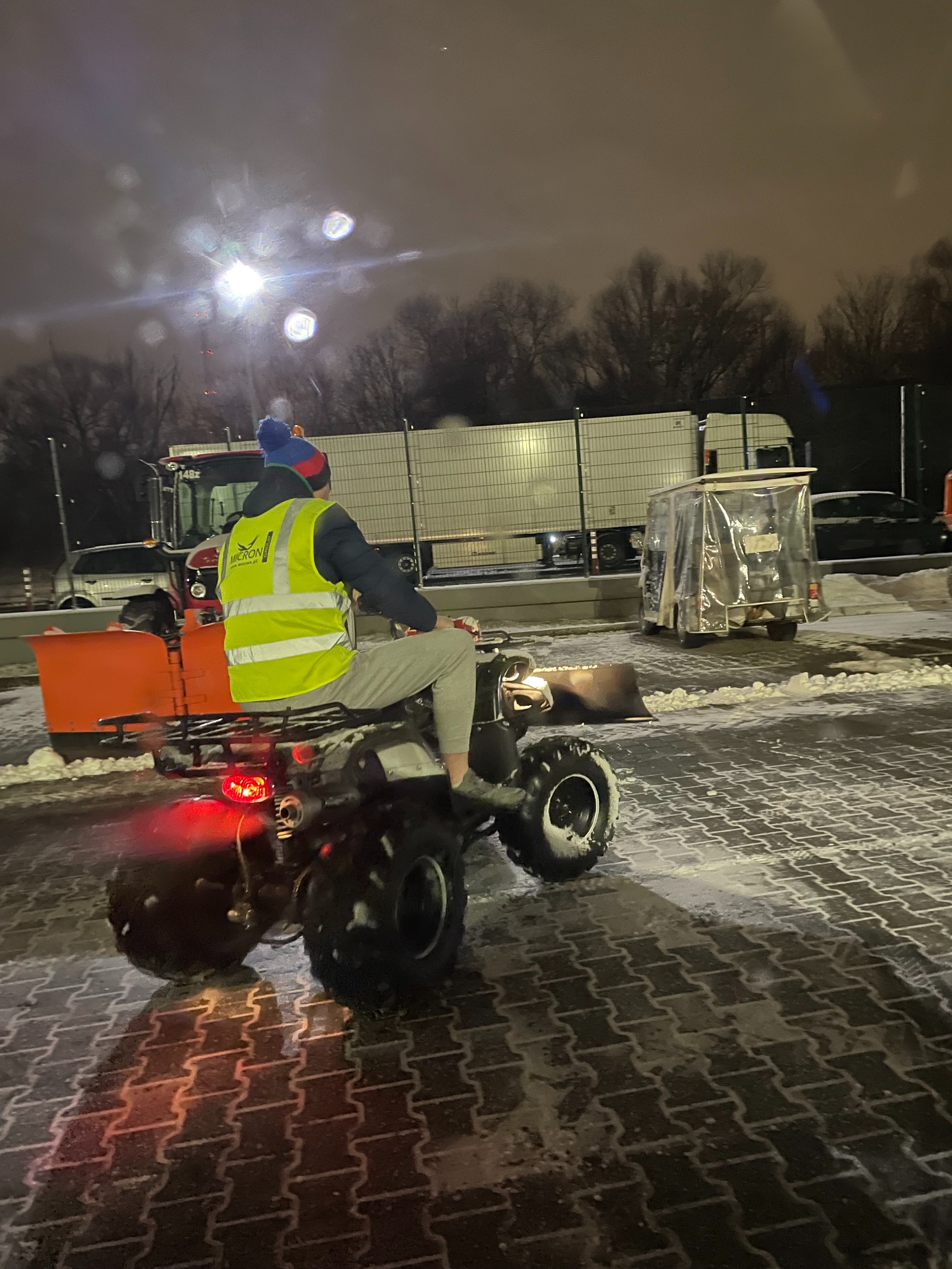Mężczyzna w odblaskowej kamizelce i czapce z pomponem siedzi na quadzie z pługiem do odśnieżania na brukowanym parkingu w nocy, w tle zaparkowane ciężarówki i drzewa.