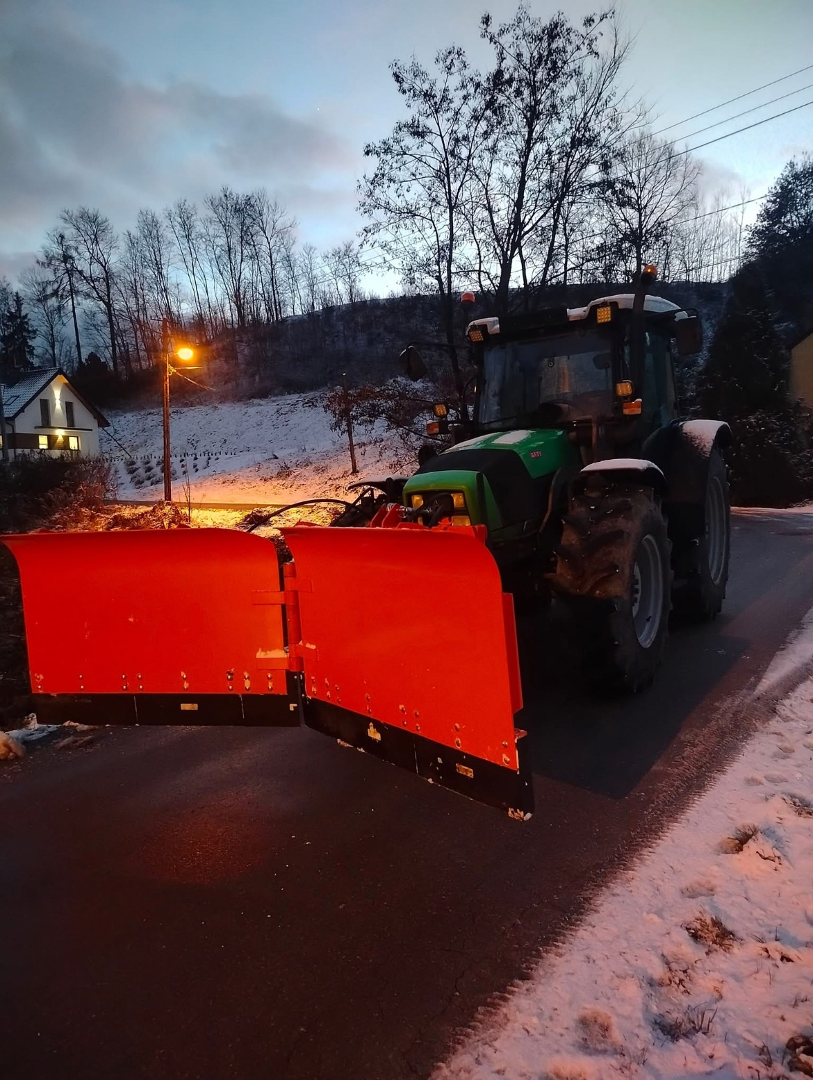 Zielony traktor z zamontowanym czerwonym pługiem do śniegu na ośnieżonej drodze o zmierzchu, w tle dom i drzewa.
