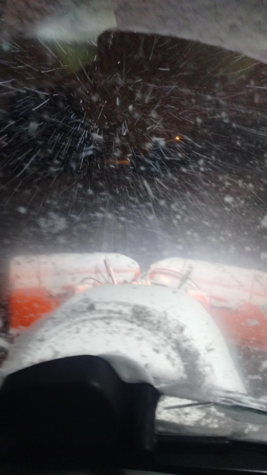 Widok zza szyby na pracującą odśnieżarkę wirnikową w trakcie intensywnych opadów śniegu, z widocznymi płatkami śniegu i elementami pomarańczowej obudowy maszyny.