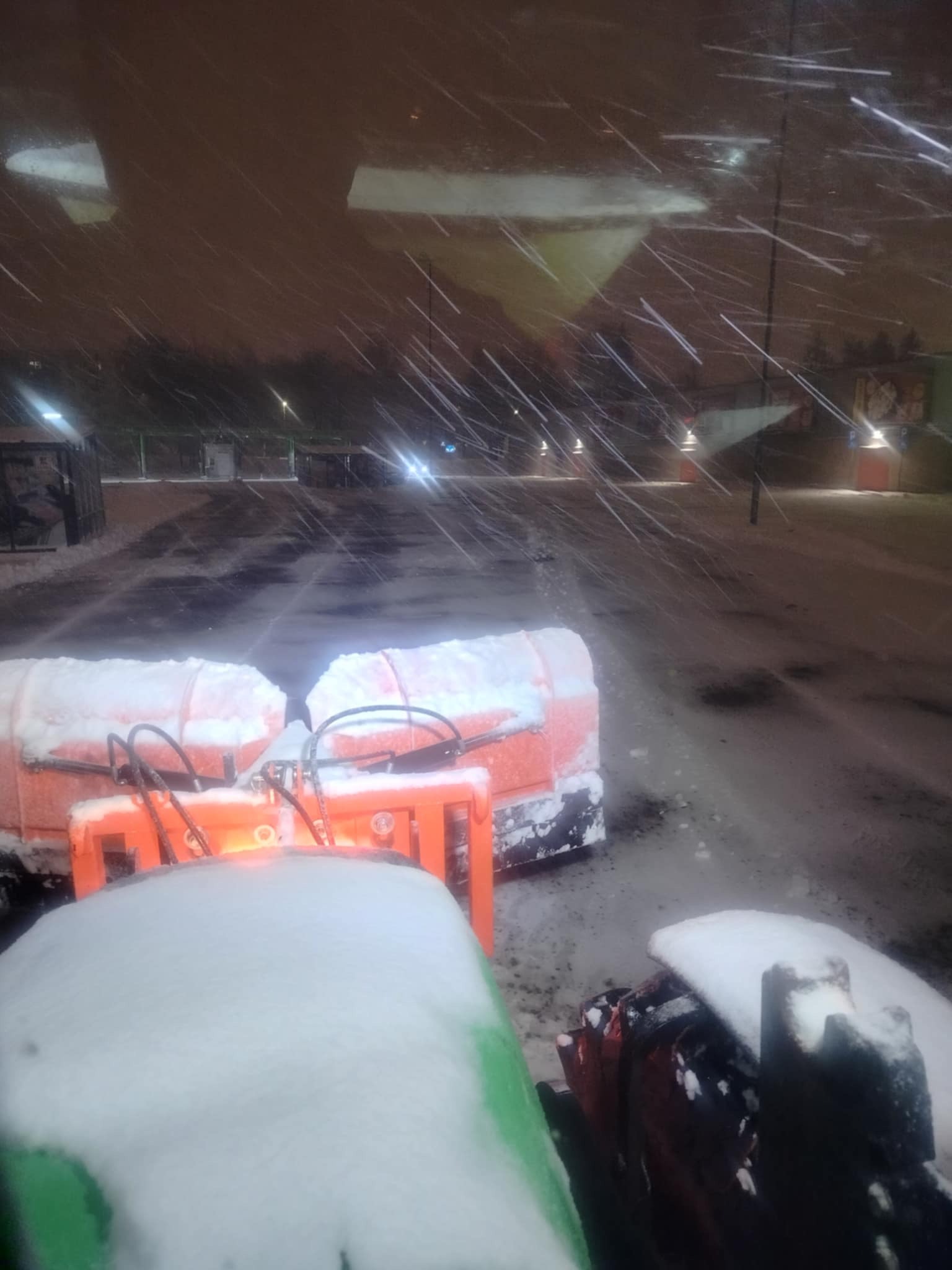 Widok z kabiny pługu śnieżnego podczas intensywnych opadów śniegu, pomarańczowe elementy pługu pokryte grubą warstwą śniegu, w tle oświetlony parking.