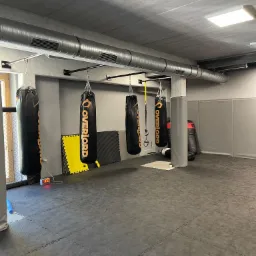 Wnętrze sali treningowej z czterema czarnymi workami bokserskimi marki OVERLORD zwisającymi z sufitu, żółtymi puzzlami podłogowymi i szarą matą podłogową.