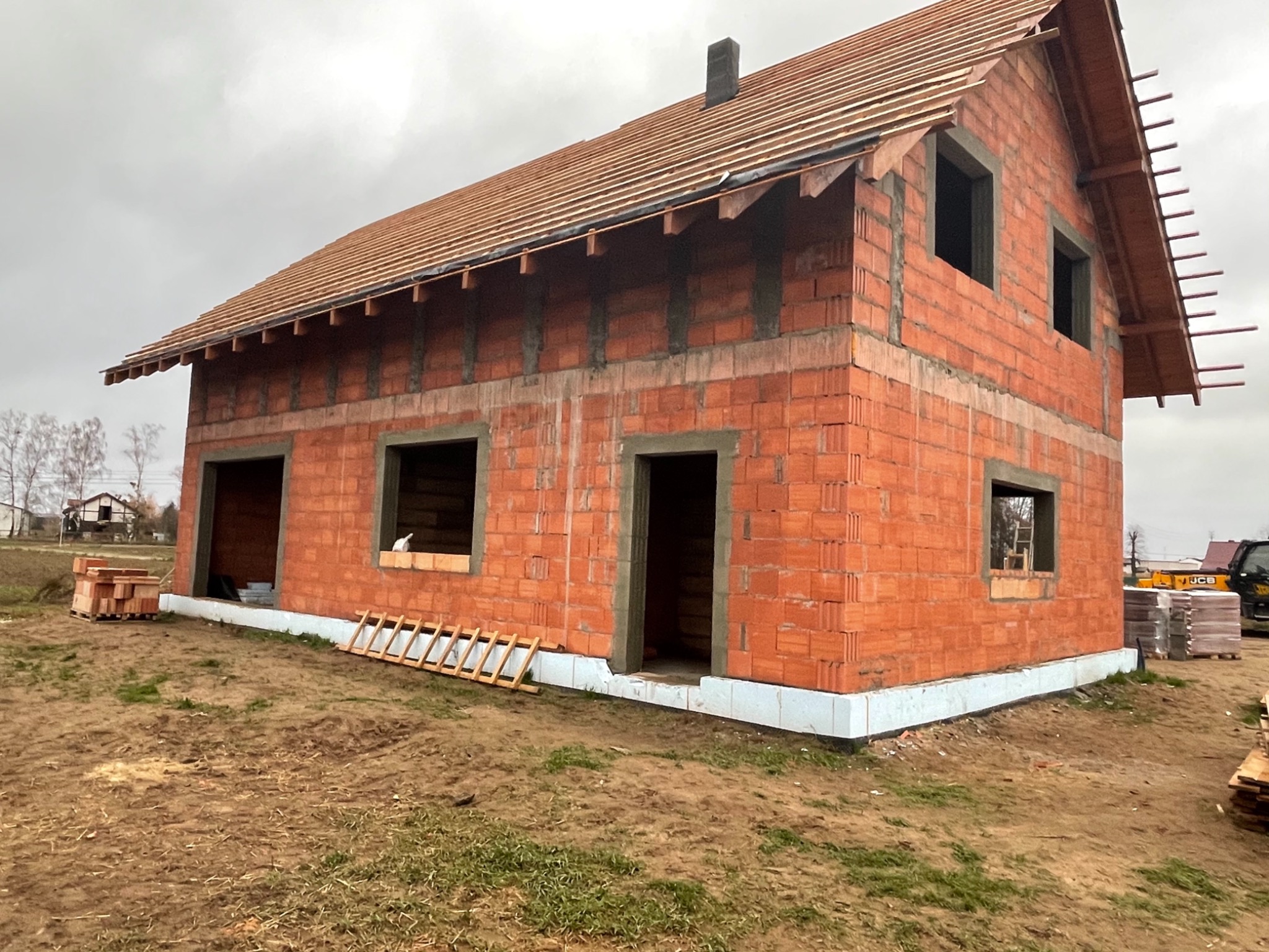 Surowy stan domu z czerwonej cegły z widoczną izolacją fundamentów, drewnianą konstrukcją dachu i stosem cegieł obok, na tle pochmurnego nieba.