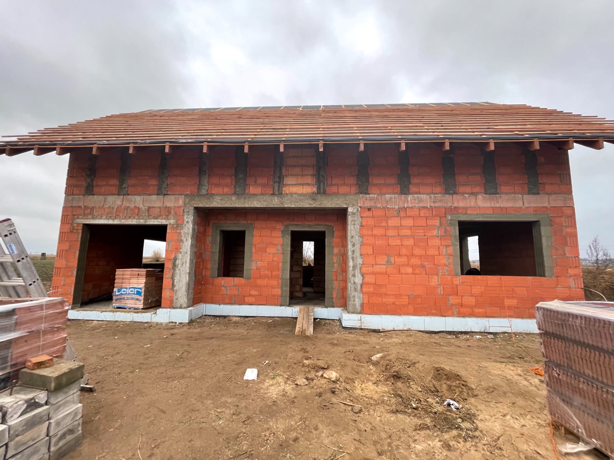 Surowy stan domu z czerwonej cegły z widocznymi otworami okiennymi i drzwiowymi, częściowo zadaszony, z paletami cegieł na pierwszym planie i drabiną opartą o ścianę.