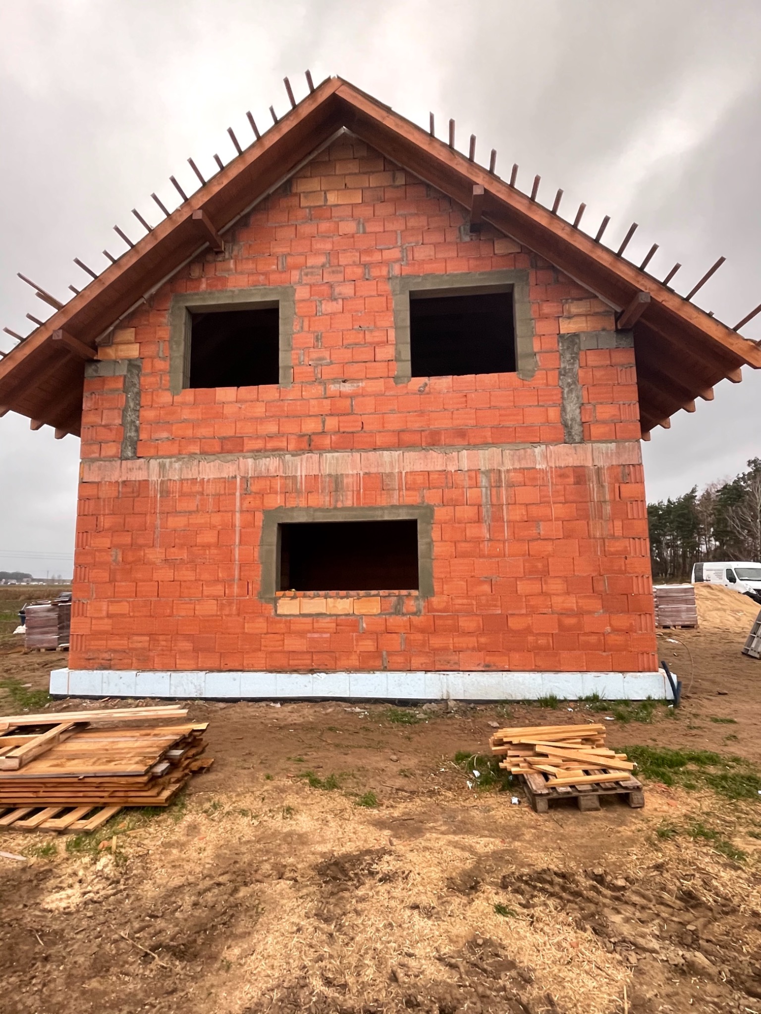 Surowy stan domu z czerwonej cegły ceramicznej z widoczną konstrukcją dachu, trzema otworami okiennymi i izolacją fundamentów, na placu budowy z paletami i ziemią.