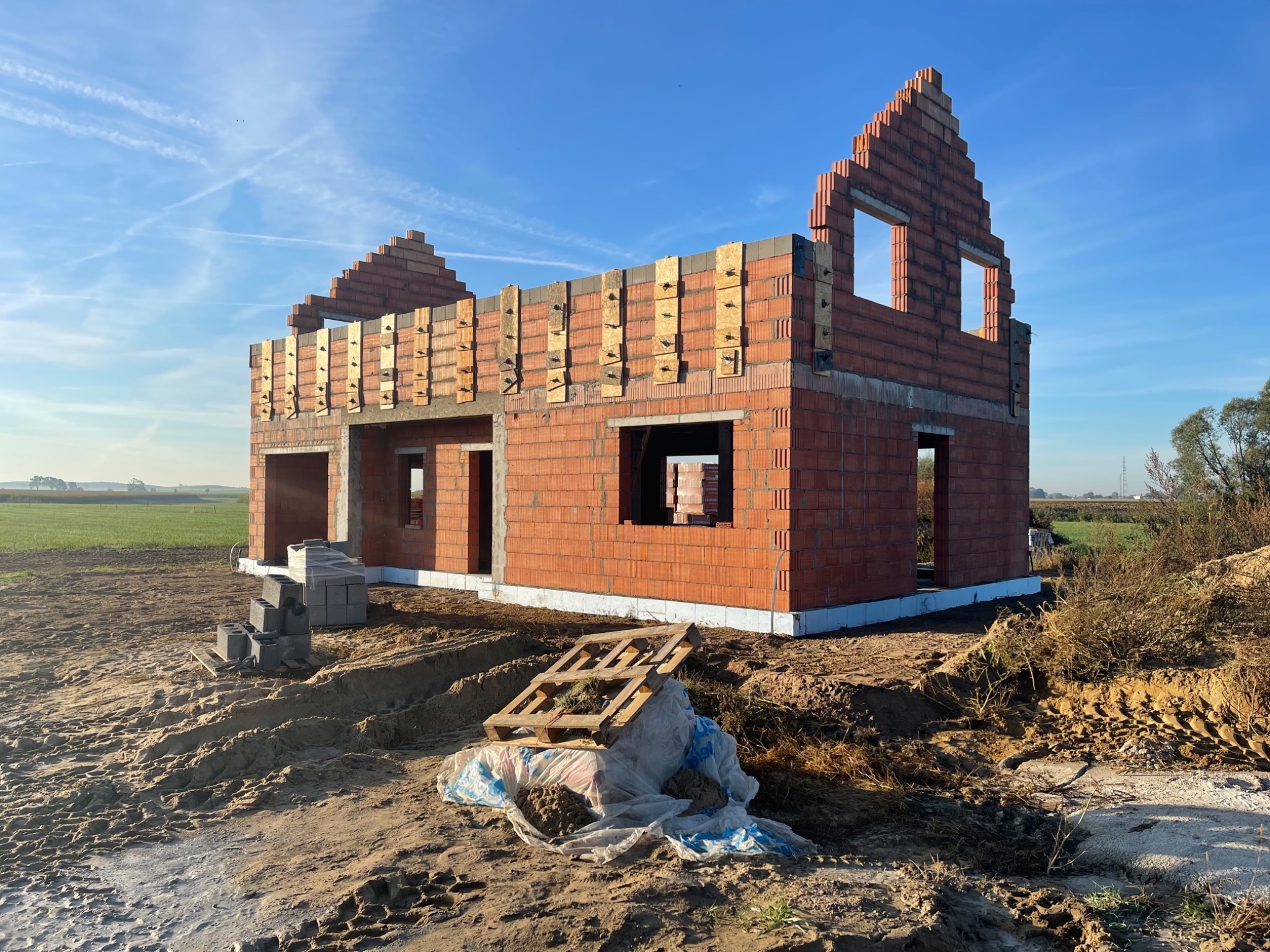 Wznoszony budynek z czerwonej cegły z widocznymi drewnianymi elementami mocującymi, na etapie budowy ścian i szczytów, z paletą leżącą na ziemi i stertą bloczków obok budynku, na tle pola...