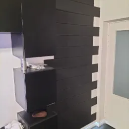 Nowoczesna aranżacja ściany z czarnymi, poziomymi panelami i podświetleniem LED u dołu, sąsiadująca z białymi szafkami wiszącymi i drzwiami.