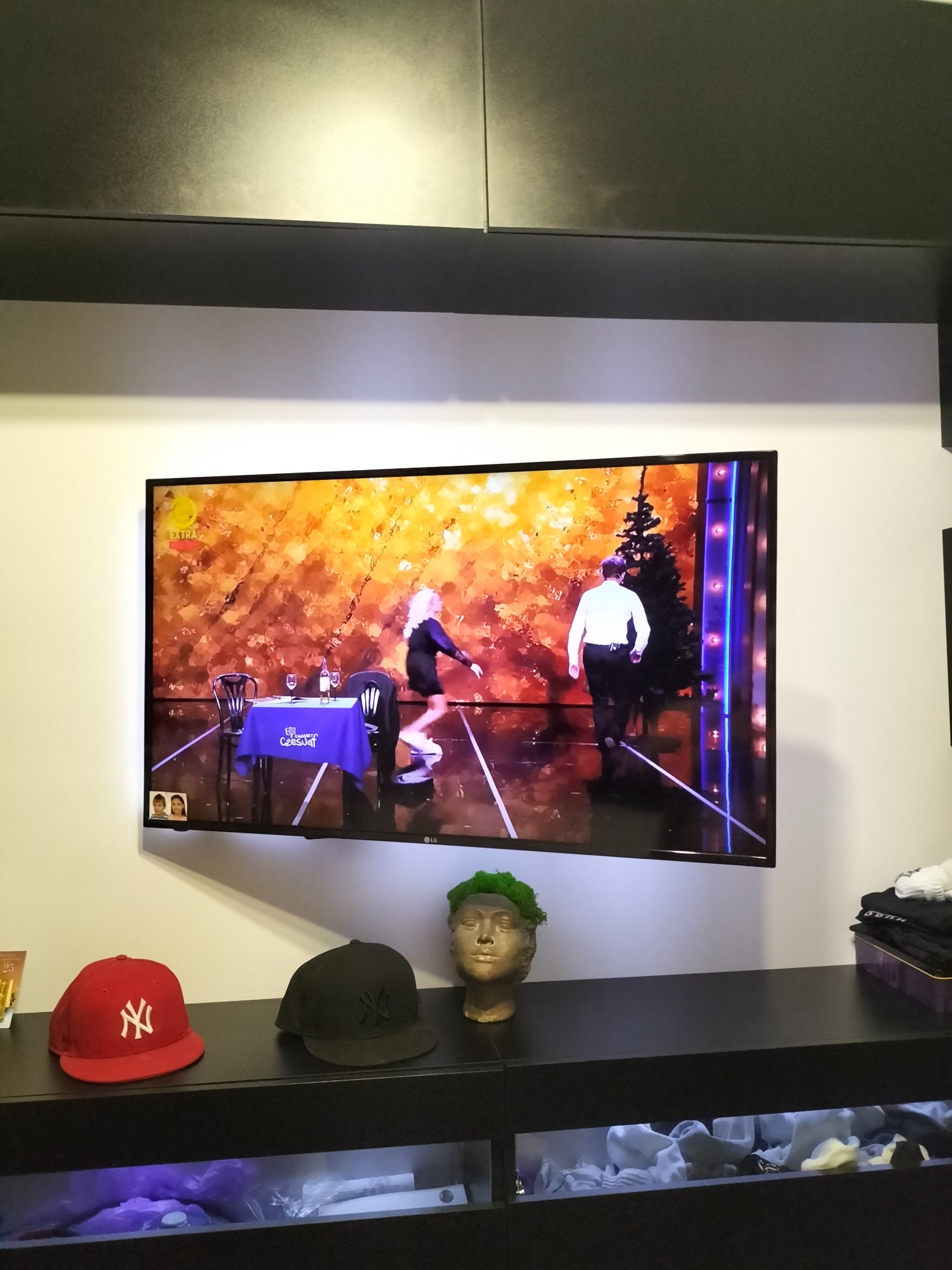 Telewizor LCD zawieszony na ścianie, wyświetlający scenę z programu rozrywkowego z udziałem kobiety i mężczyzny przy stole, poniżej szafka RTV z czapkami New York Yankees i popiersiem z mchu.