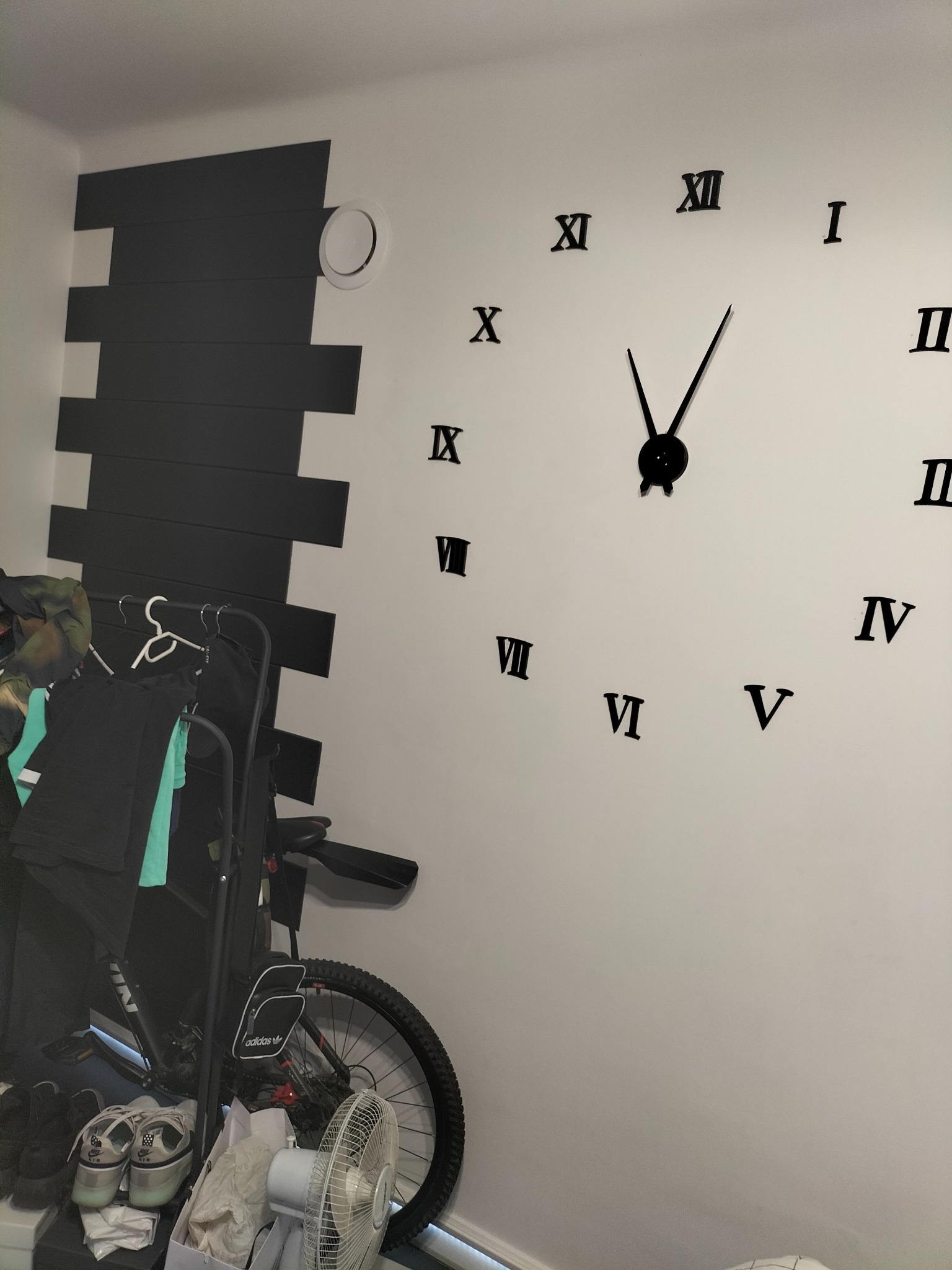 Ściana z dekoracyjnymi czarnymi panelami i dużym zegarem z rzymskimi cyframi, obok wieszak na ubrania i rower.