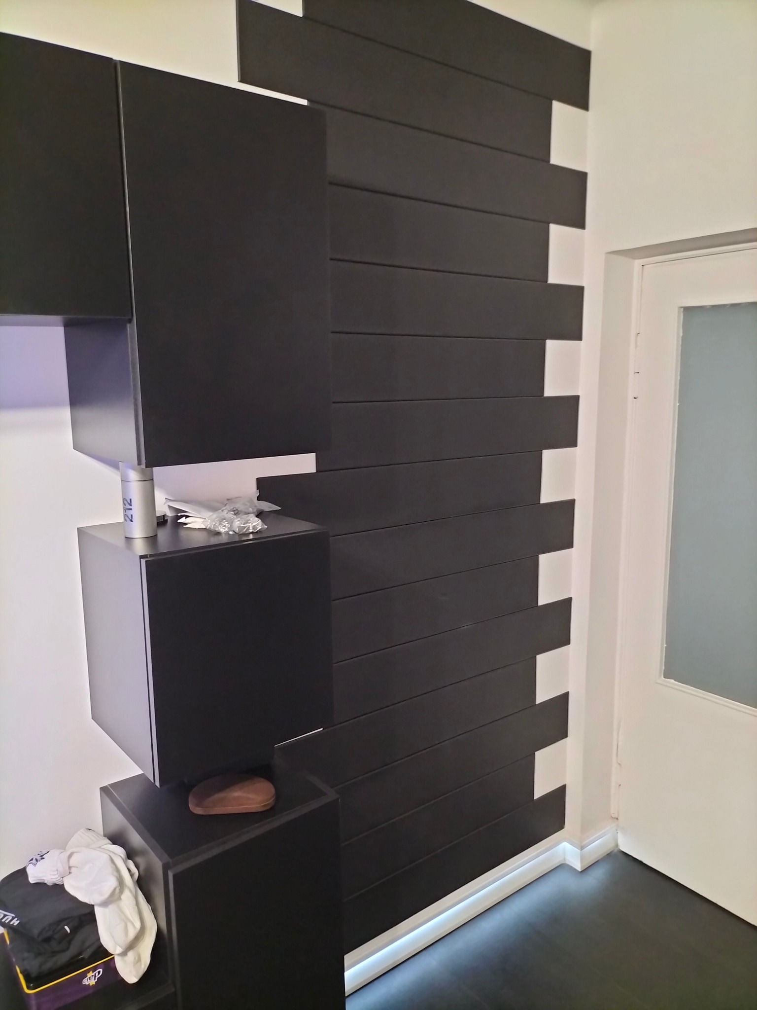 Nowoczesna aranżacja ściany z czarnymi, poziomymi panelami i podświetleniem LED u dołu, sąsiadująca z białymi szafkami wiszącymi i drzwiami.