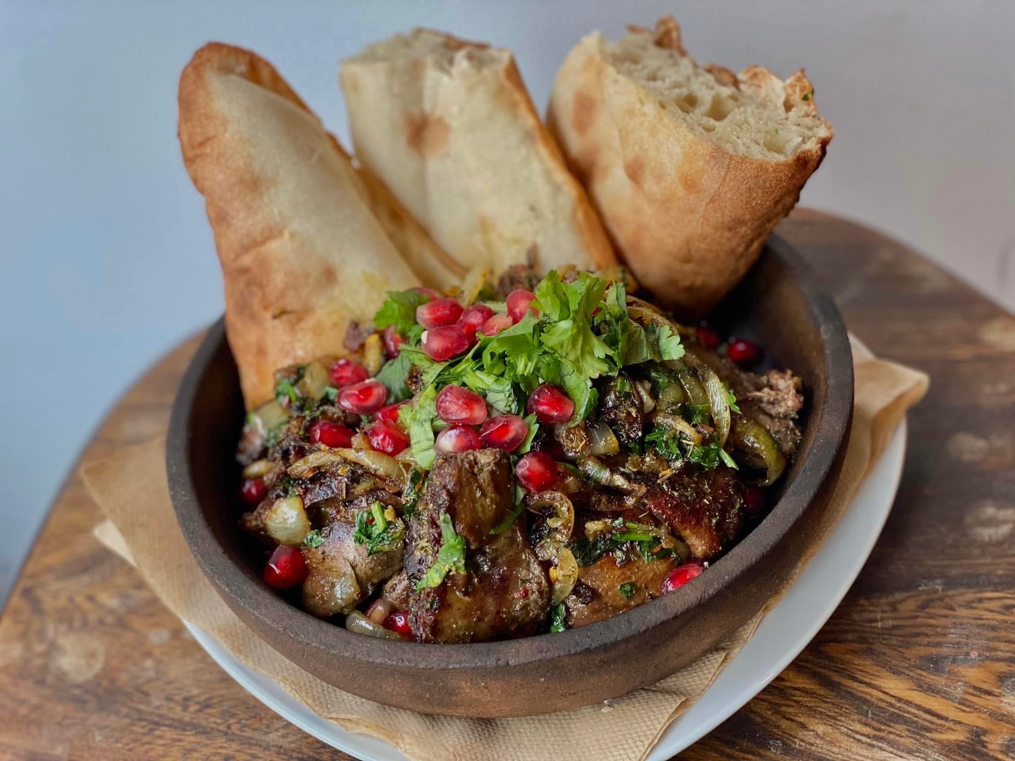 Danie mięsne w glinianym naczyniu, udekorowane granatem i kolendrą, podane z chlebem lawasz na drewnianym stole.