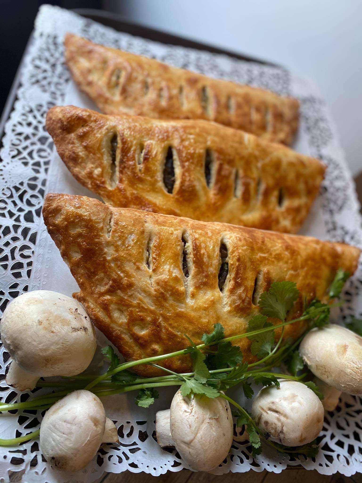 Trzy złociste paszteciki z farszem, ułożone na ozdobnej serwetce, udekorowane świeżą kolendrą i surowymi pieczarkami.