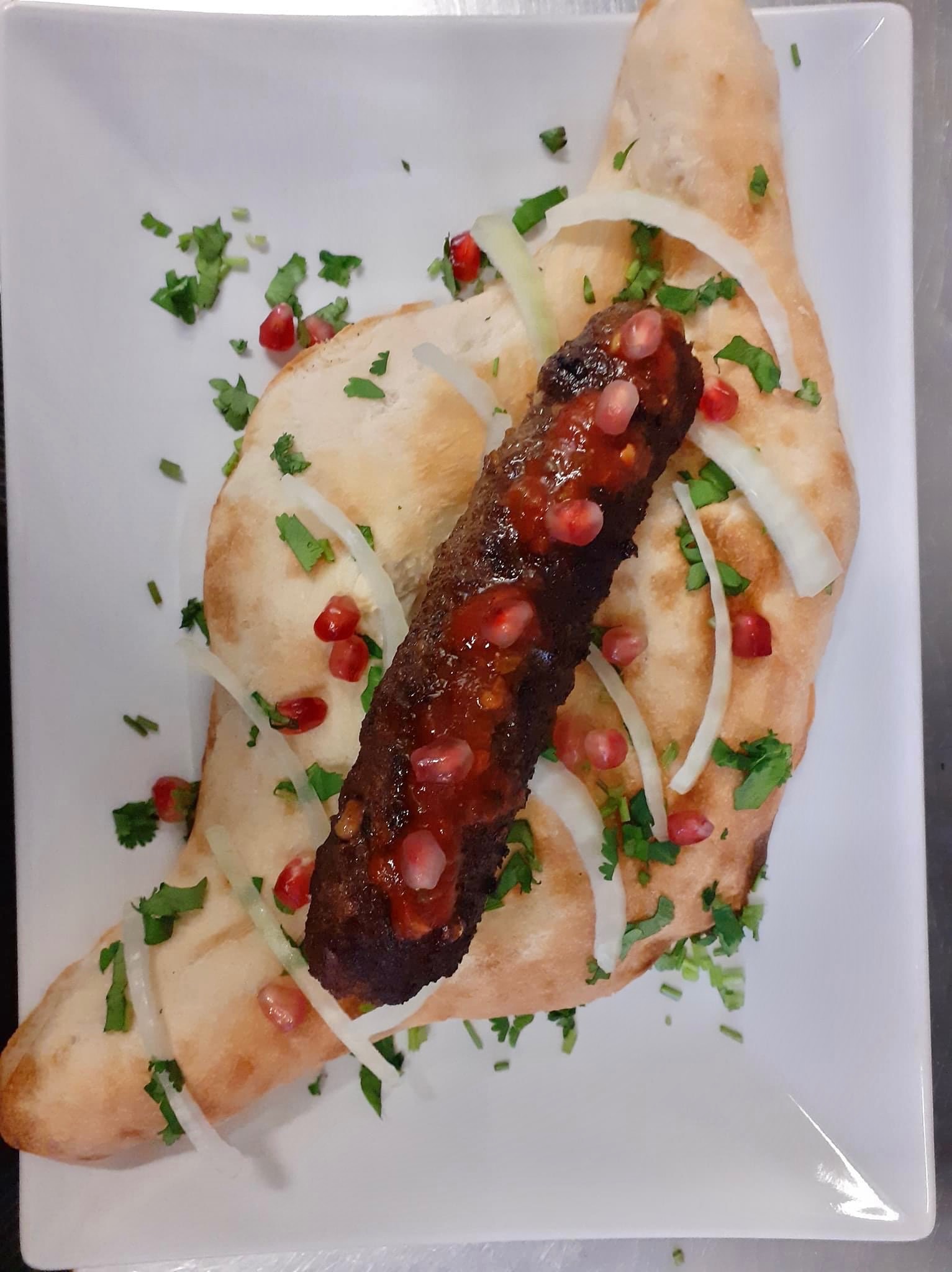 Kebab z granatem na chlebie lawasz, udekorowany cebulą i kolendrą, podany na białym kwadratowym talerzu.