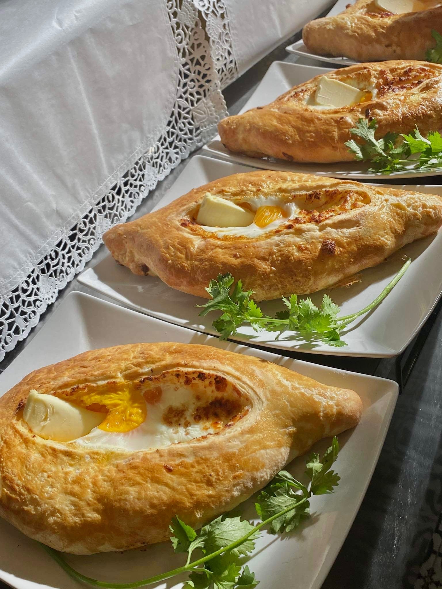 Trzy porcje chaczapuri adżarskiego na białych talerzach, udekorowane świeżą kolendrą i ułożone na stole przykrytym białym obrusem z koronkowym wykończeniem.
