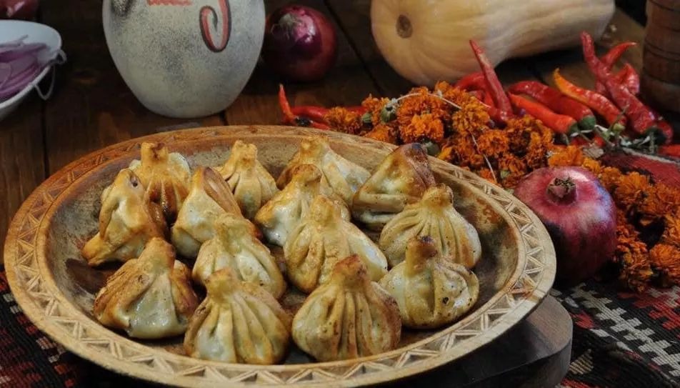 Gruzińskie pierożki chinkali ułożone na drewnianym talerzu, otoczone dekoracjami z granatu, papryczek chili i pomarańczowych kwiatów, z dynią i cebulą w tle.