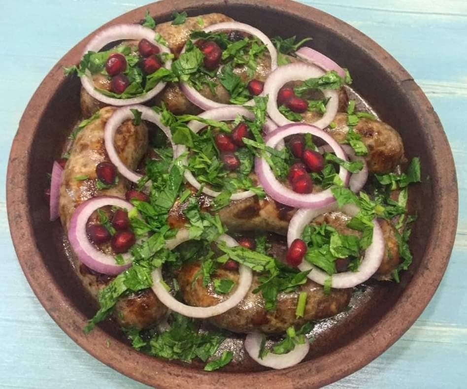 Grillowane kiełbaski w glinianej misce, udekorowane świeżą kolendrą, krążkami czerwonej cebuli i pestkami granatu.