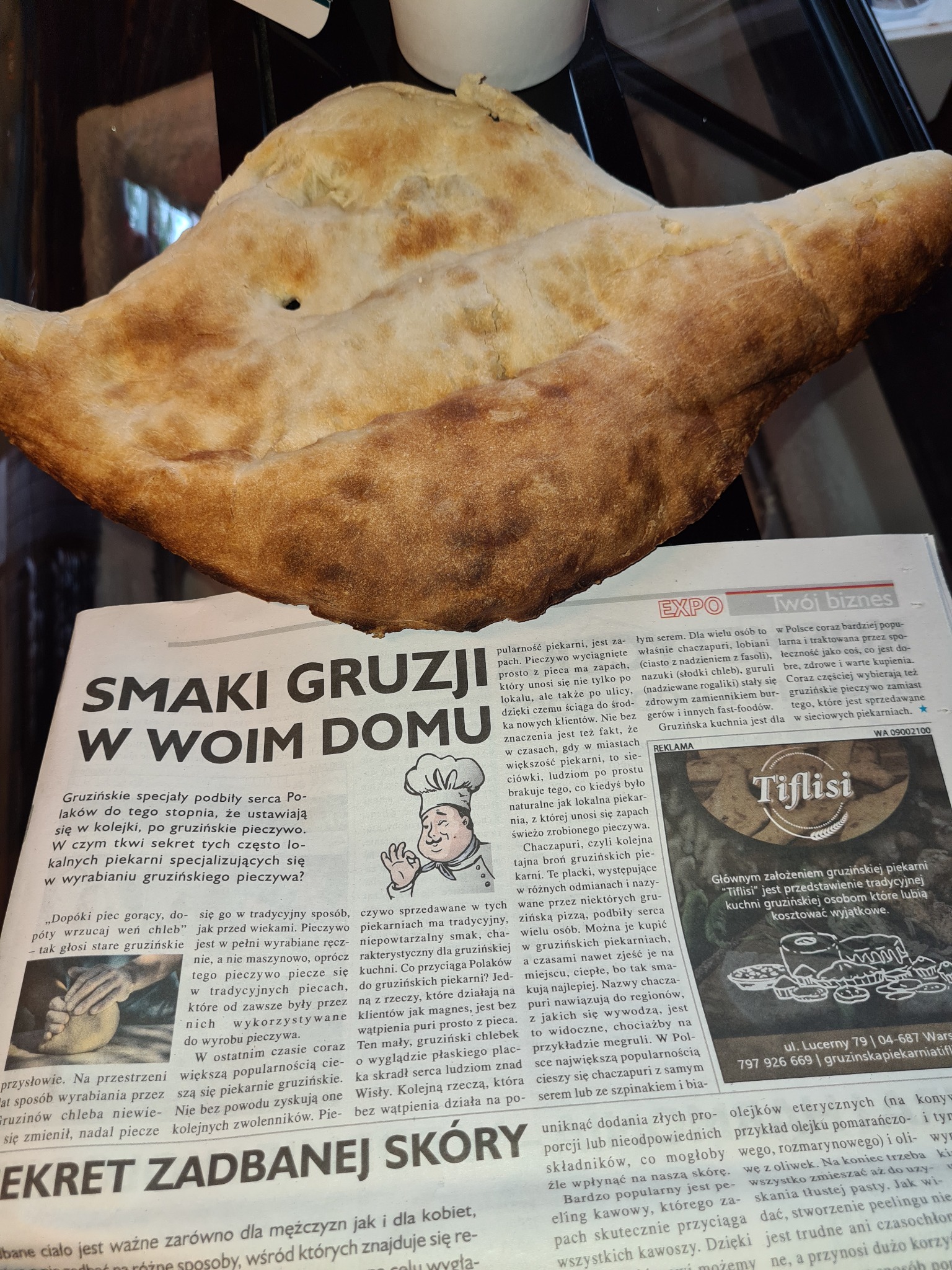 Duży chleb lawasz leży na gazecie z artykułem o gruzińskich smakach, w tle niewyraźne elementy wyposażenia.