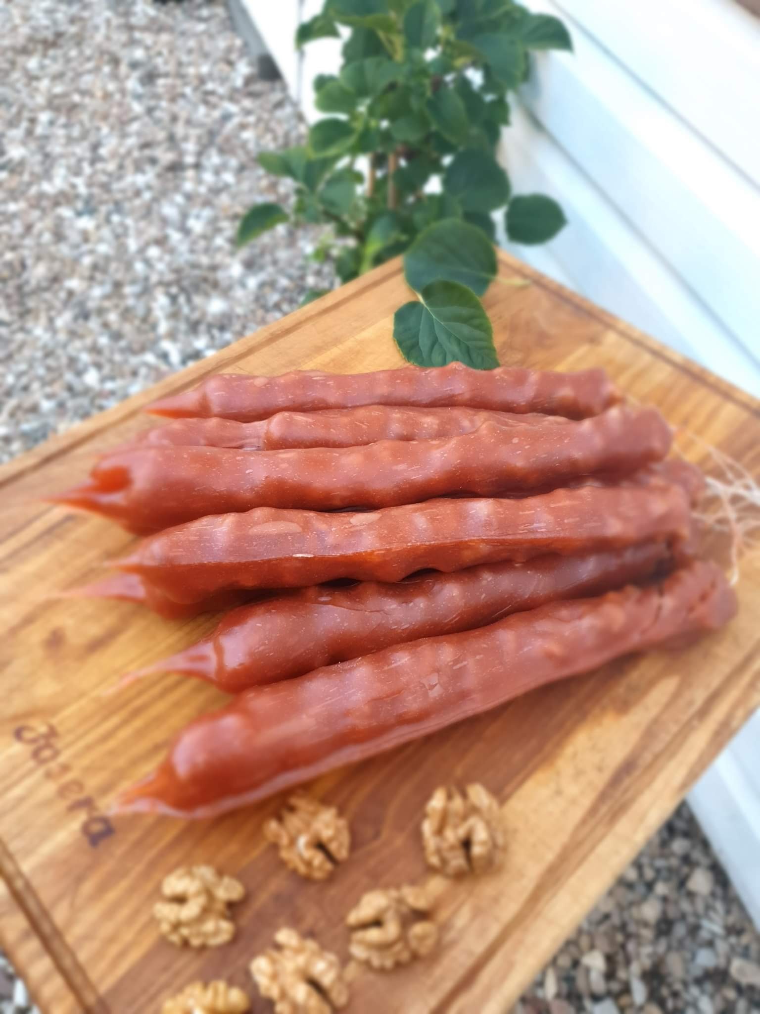 Warszawski przysmak: gruzińskie czurczchele ułożone na drewnianej desce, otoczone połówkami orzechów włoskich, z rośliną w tle.