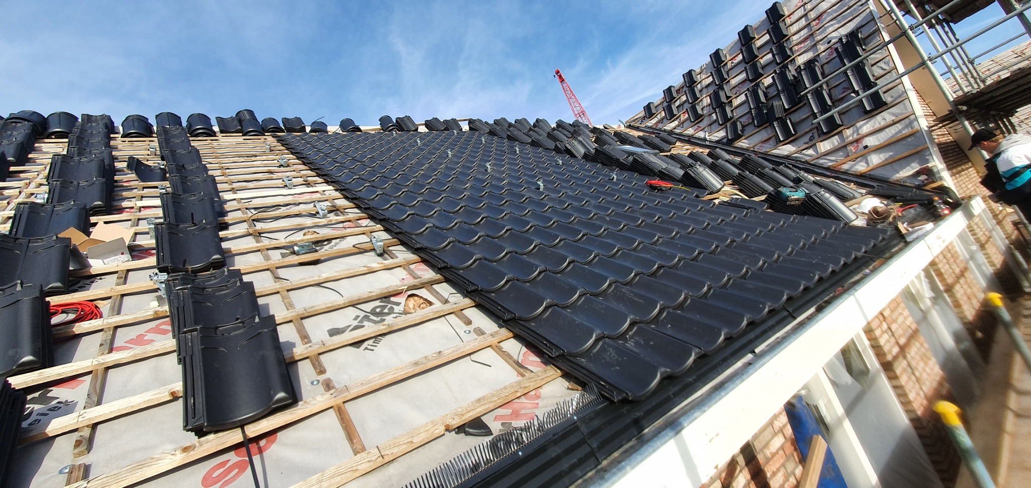 Układanie czarnej dachówki na nowym dachu, widoczne łaty, membrana dachowa, narzędzia i rusztowanie w tle.
