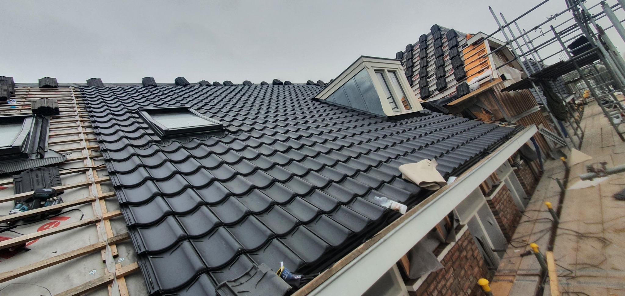 Częściowo pokryty czarną dachówką dach domu z oknami dachowymi i lukarną w trakcie prac dekarskich, widoczne rusztowania i przygotowane elementy do dalszego krycia.