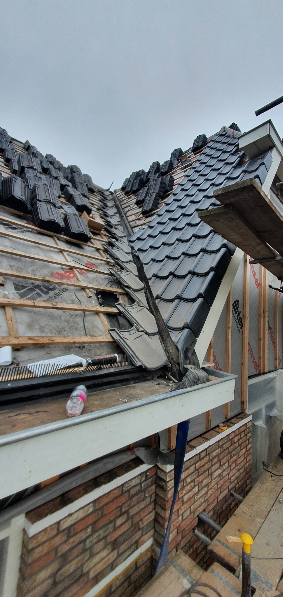 Montaż czarnej dachówki ceramicznej na skośnym dachu, widoczne elementy konstrukcyjne, membrana dachowa i rynna, częściowo ukończona połać dachu.