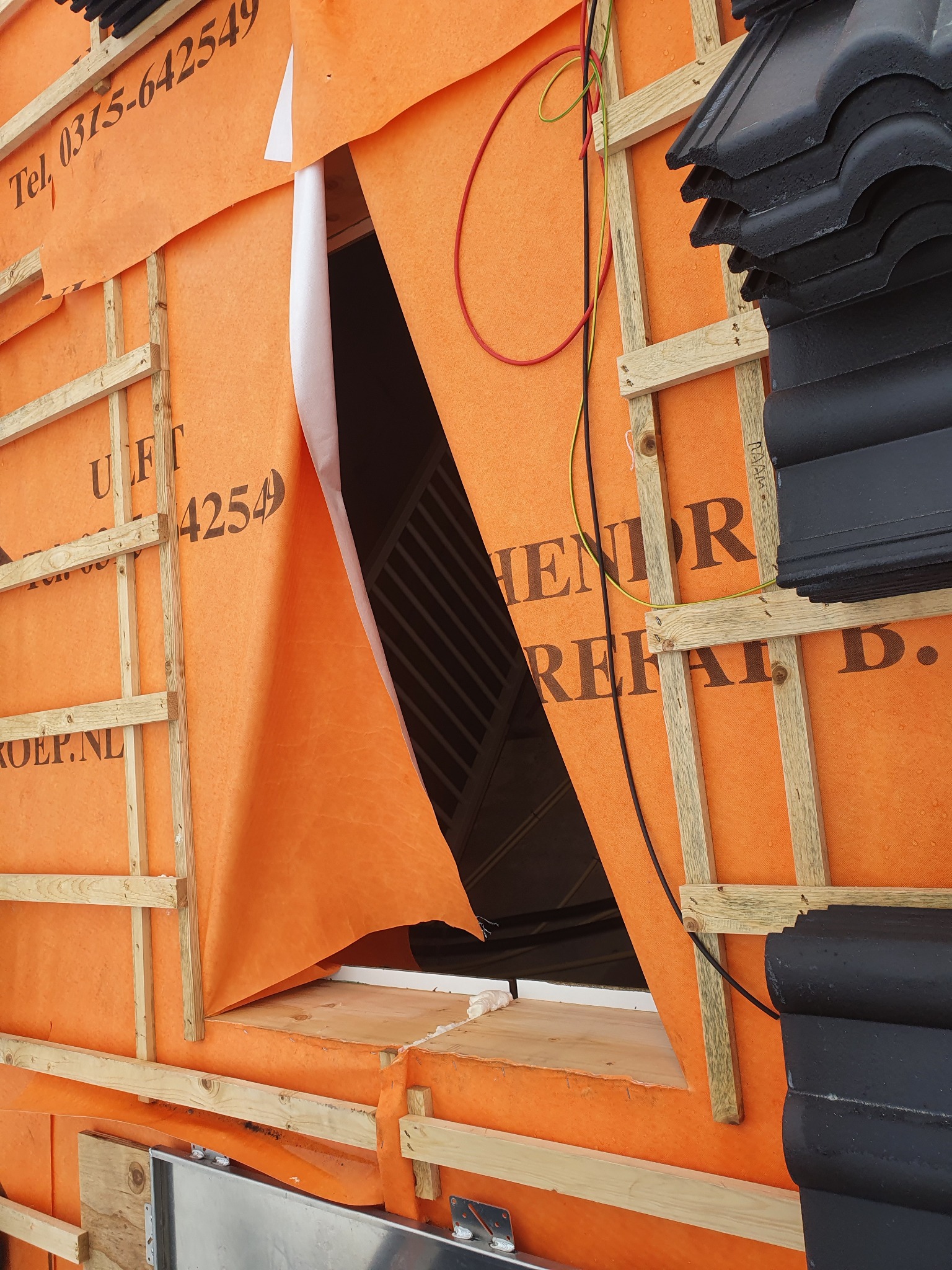 Montaż pomarańczowej membrany dachowej z widoczną konstrukcją drewnianą, fragmentem okna dachowego i stosem czarnych dachówek. Widoczne przewody elektryczne.