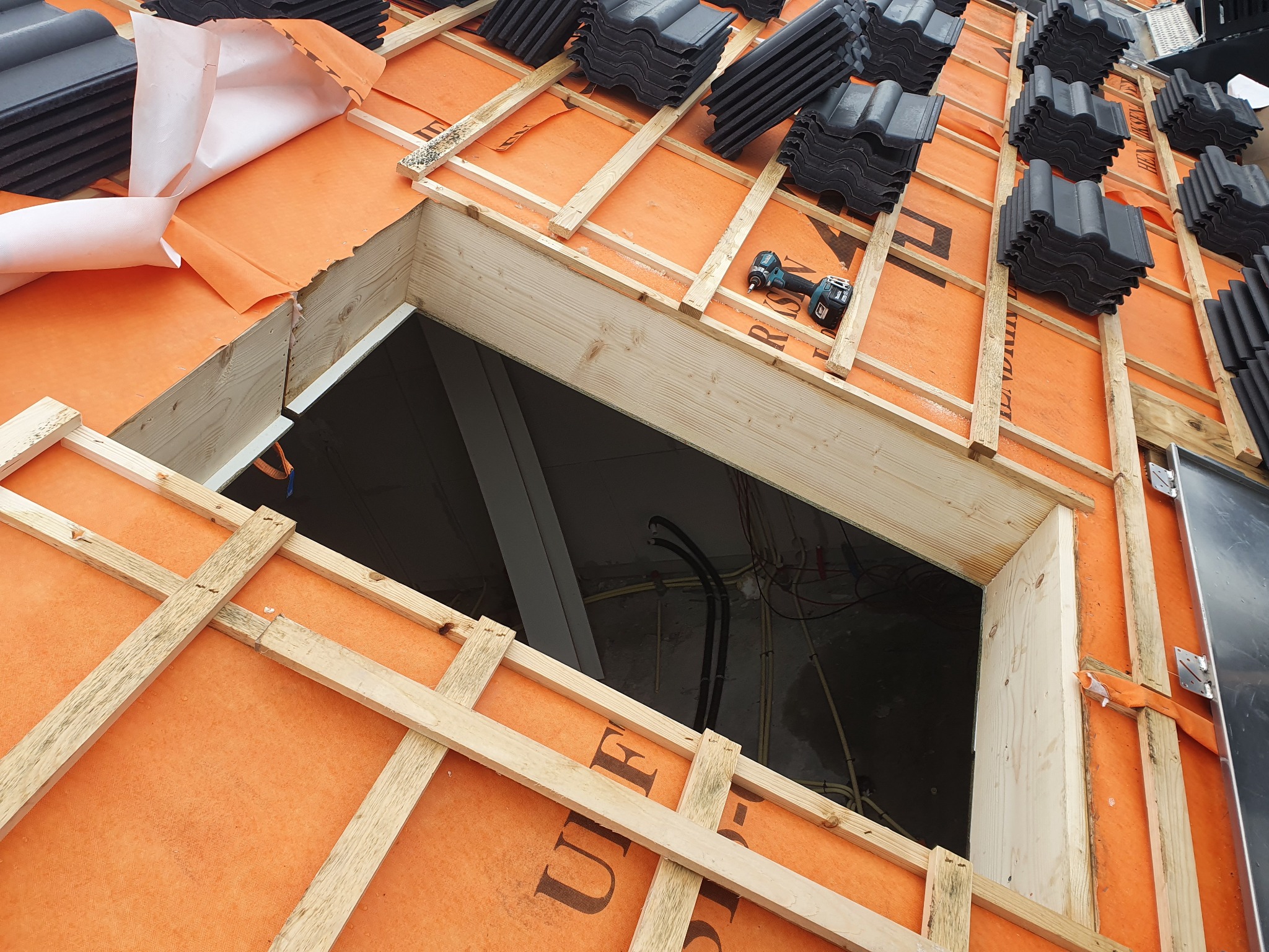 Montaż dachu: widok od wewnątrz, na pomarańczowej membranie ułożone drewniane łaty, na nich ułożone dachówki, w tle otwór okienny i wkrętarka.