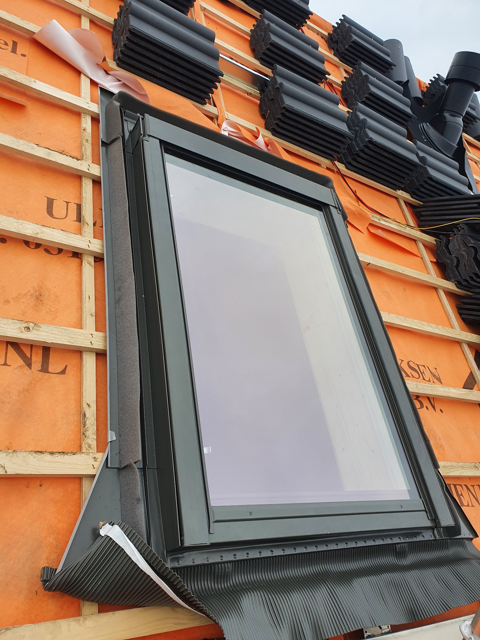 Montaż okna dachowego na drewnianej konstrukcji z pomarańczową membraną, widoczne czarne dachówki w tle i uszczelnienia wokół okna.
