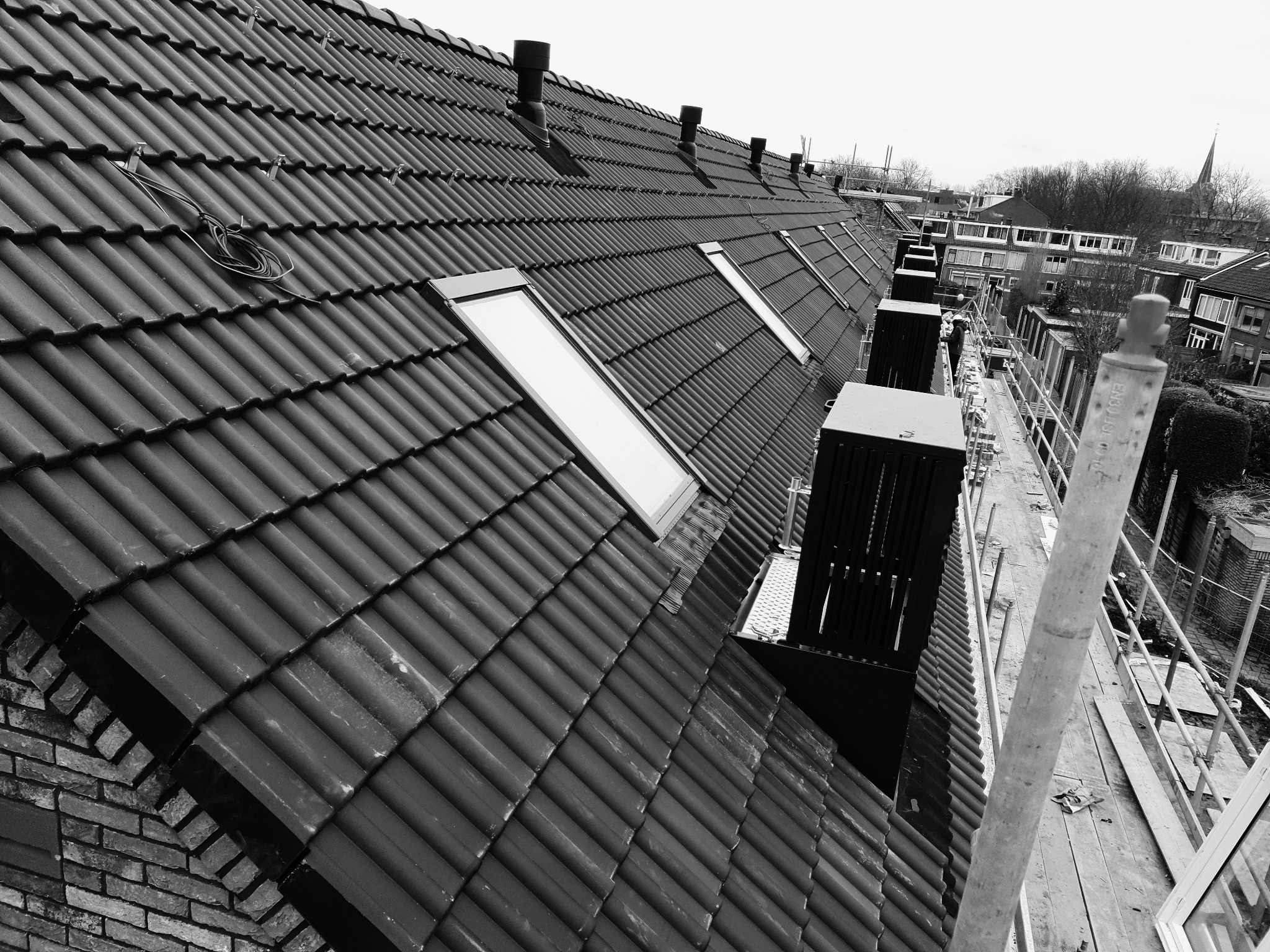 Czarno-białe zdjęcie dachu z dachówką karpiówką i oknami dachowymi w Głubczycach, widok z rusztowania, w tle budynki mieszkalne i wieża kościoła.