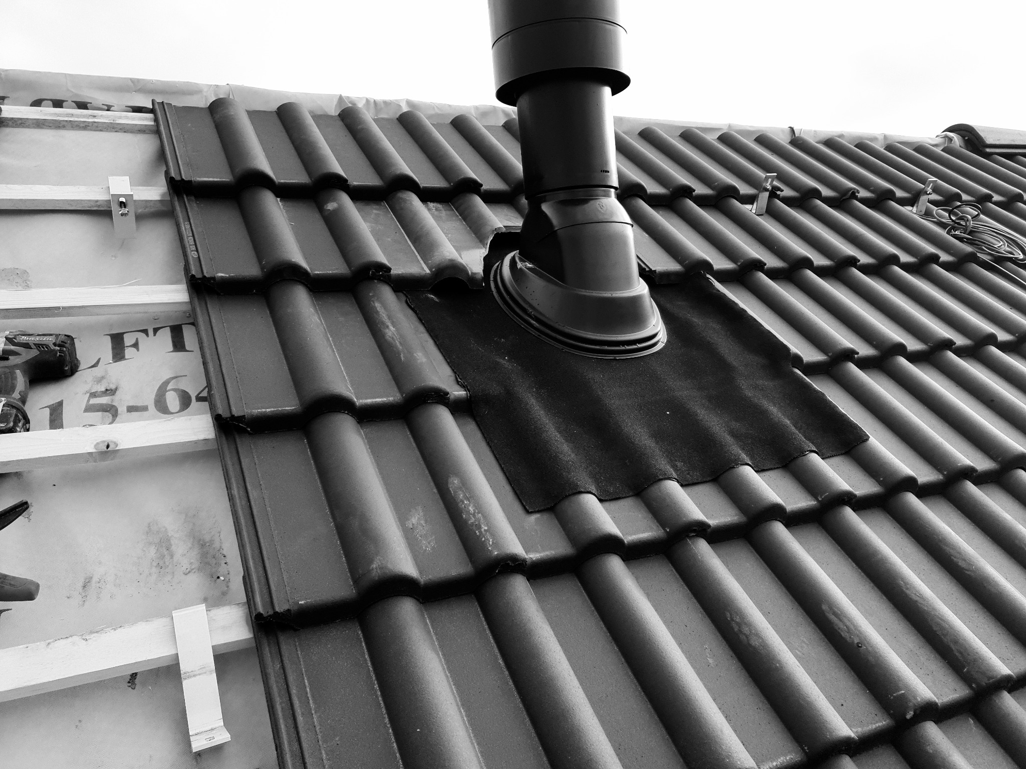 Fragment dachu z zamontowaną czarną rurą wentylacyjną i częściowo ułożoną dachówką ceramiczną na łatach, widoczne narzędzia dekarskie.