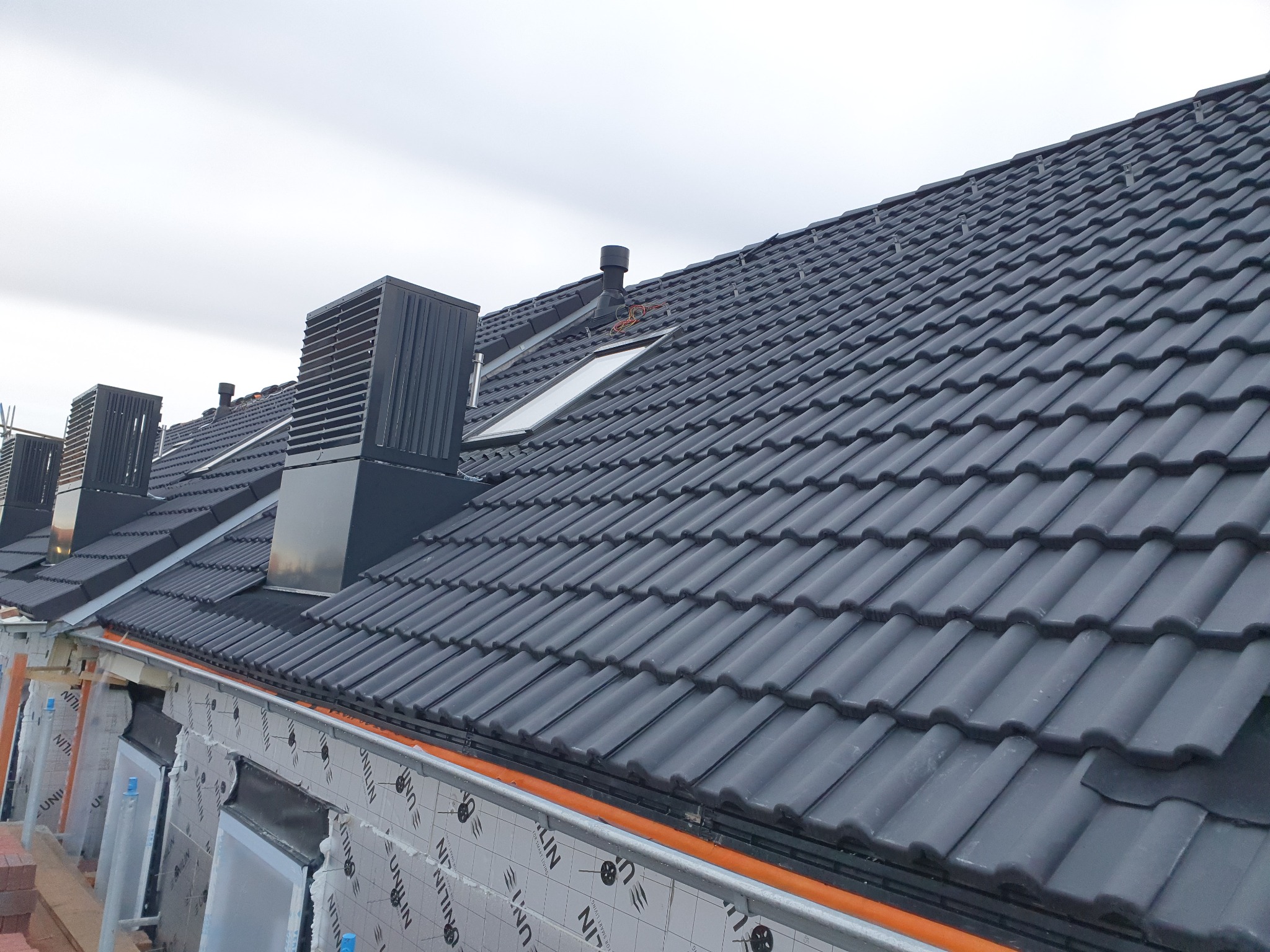 Nowy dach z czarnej dachówki ceramicznej, widoczne kominy wentylacyjne i okno dachowe, elewacja w trakcie ocieplania, zabezpieczona folią.