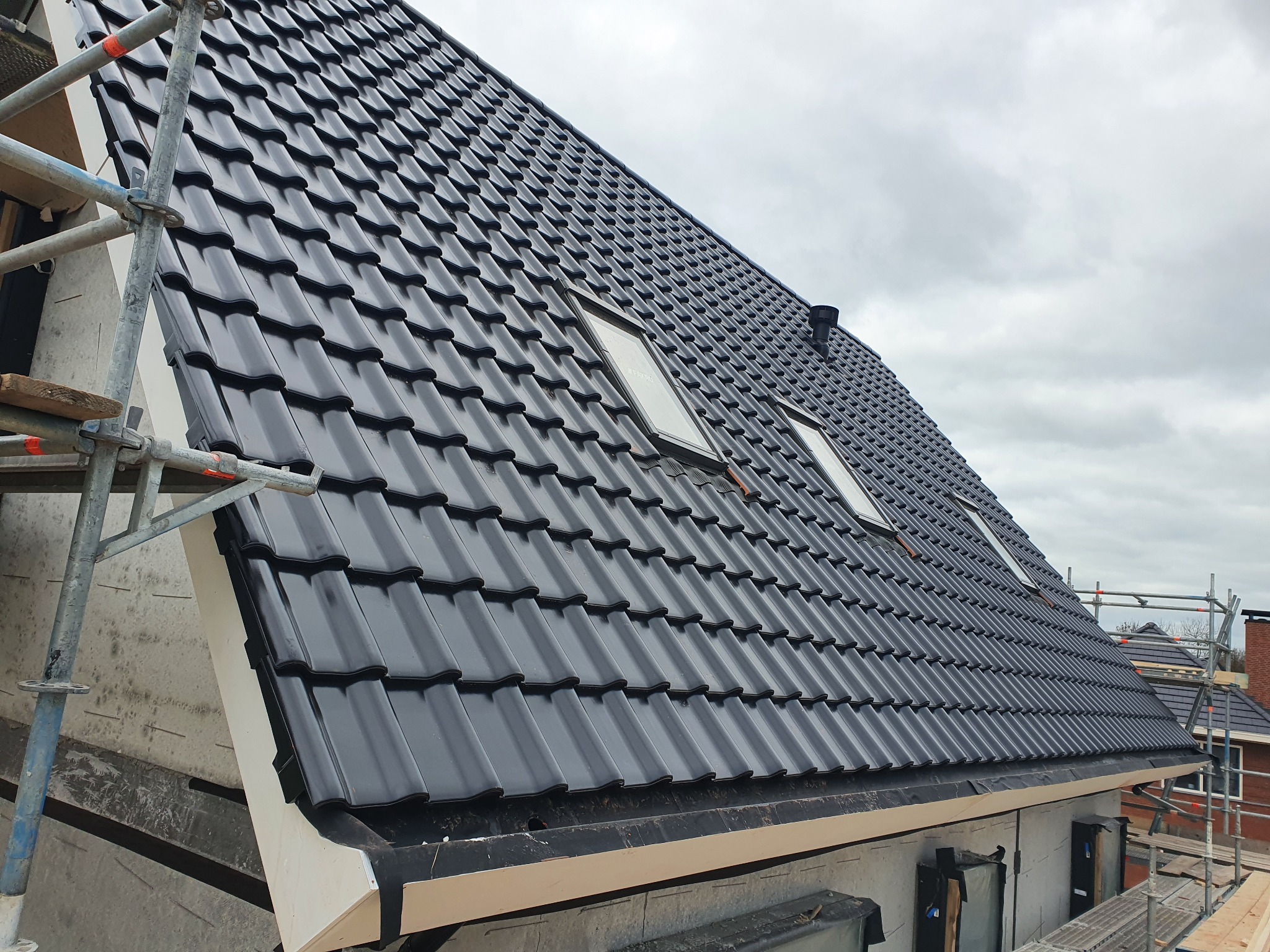 Nowy, błyszczący dach z czarnej dachówki ceramicznej z oknami dachowymi, widoczny fragment rusztowania, pochmurne niebo w tle.