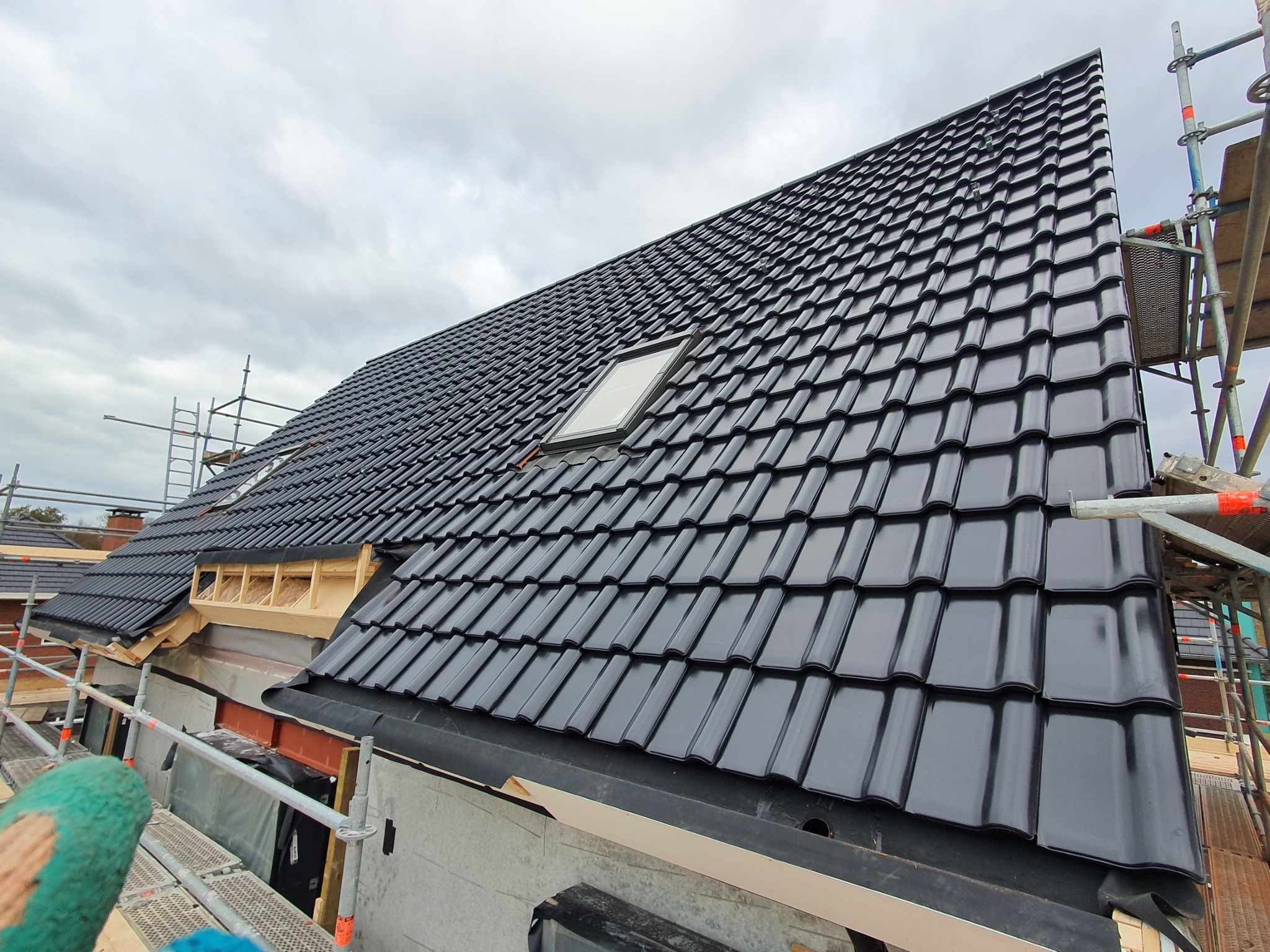 Nowy dach z czarnej dachówki ceramicznej z oknem dachowym, widoczny z rusztowania podczas montażu, pochmurne niebo w tle.