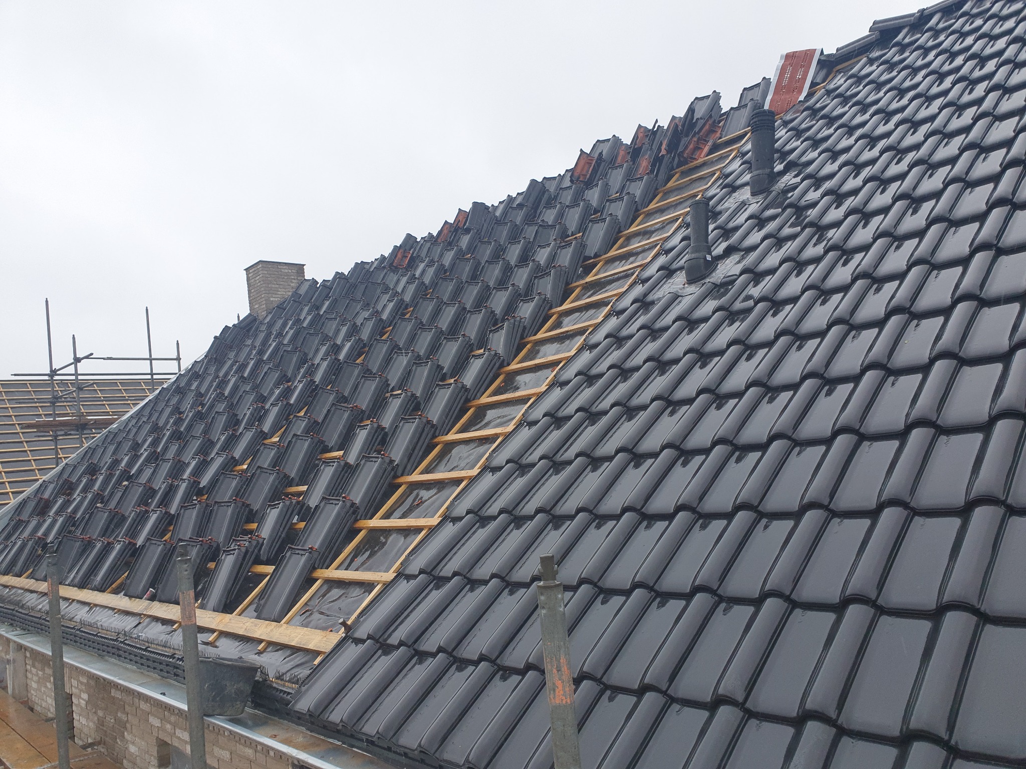 Częściowo pokryty dach z czarnej dachówki ceramicznej, widoczne łaty drewniane i komin w tle.