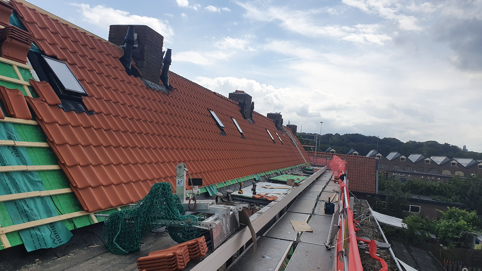 Czerwona dachówka na skośnym dachu w trakcie prac, widoczne okna dachowe, kominy, rusztowanie i narzędzia, w tle zabudowania miejskie i zieleń.
