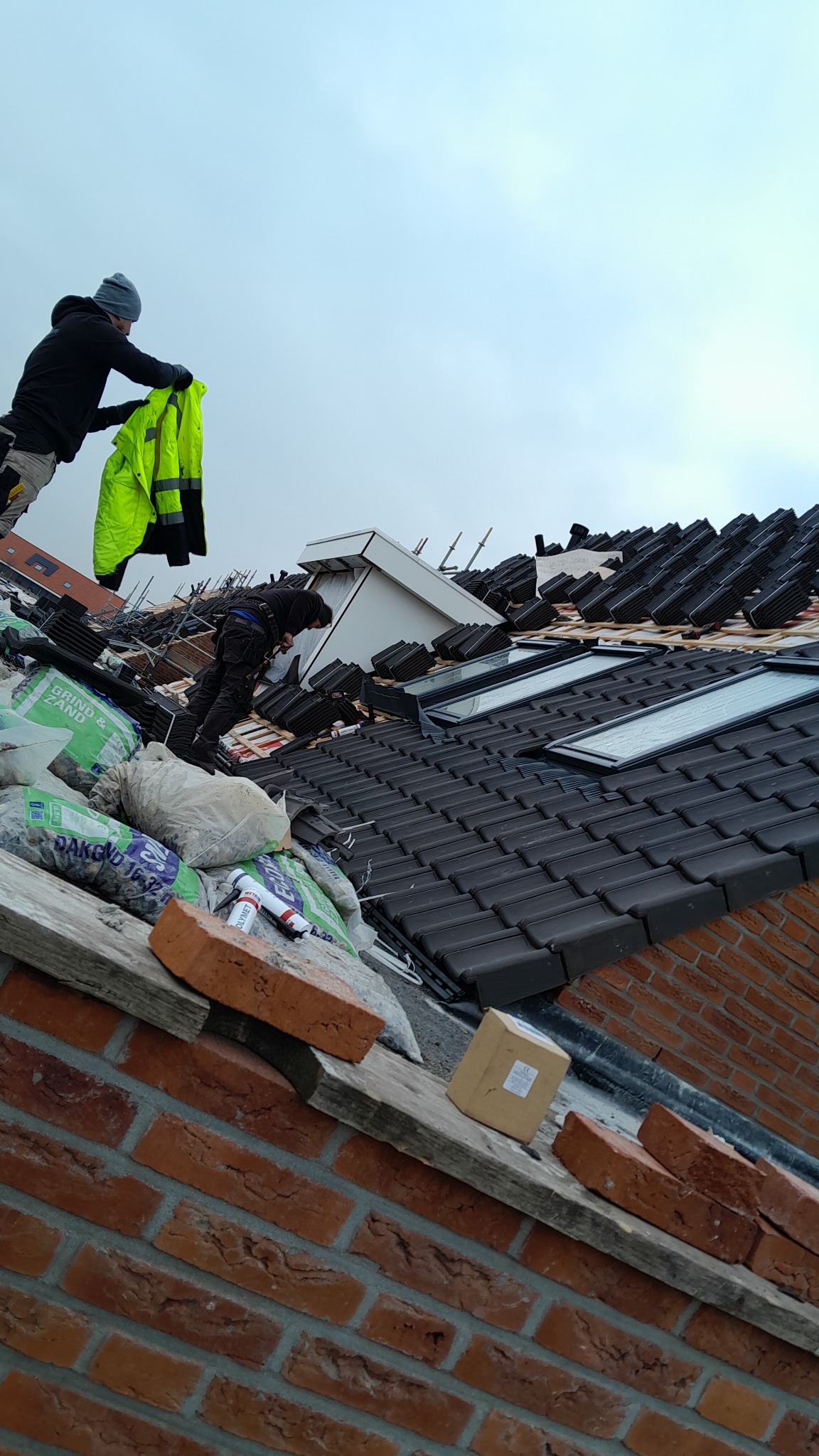Prace dekarskie w toku: dekarz w czarnej odzieży układa ciemne dachówki na dachu z oknami połaciowymi, obok worki z materiałami i cegły, drugi dekarz trzyma żółtą kurtkę roboczą.