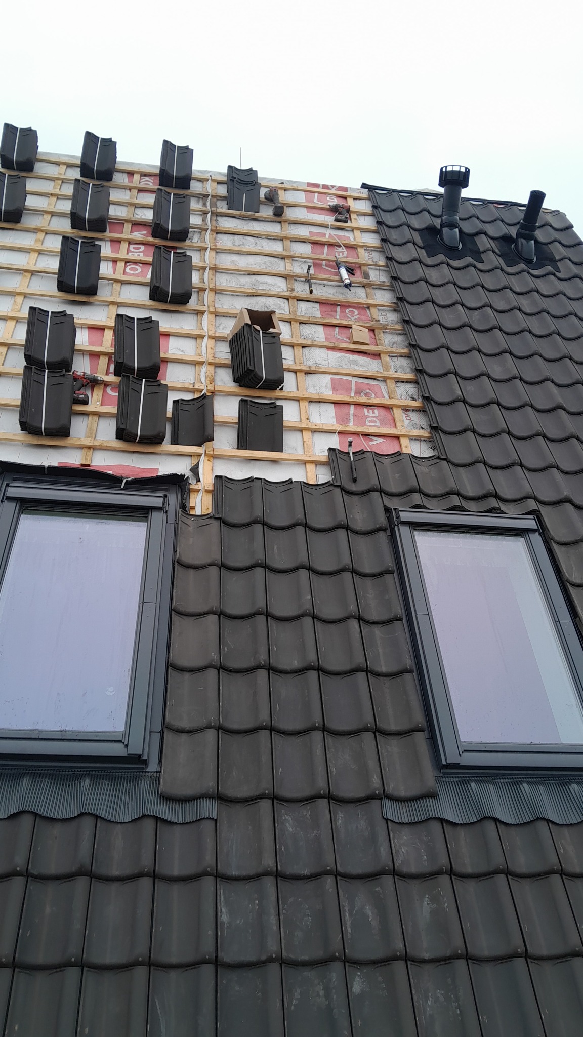 Częściowo pokryty dach z dwoma oknami dachowymi, widoczne przygotowane do ułożenia dachówki na łatach, narzędzia i materiały budowlane na dachu.