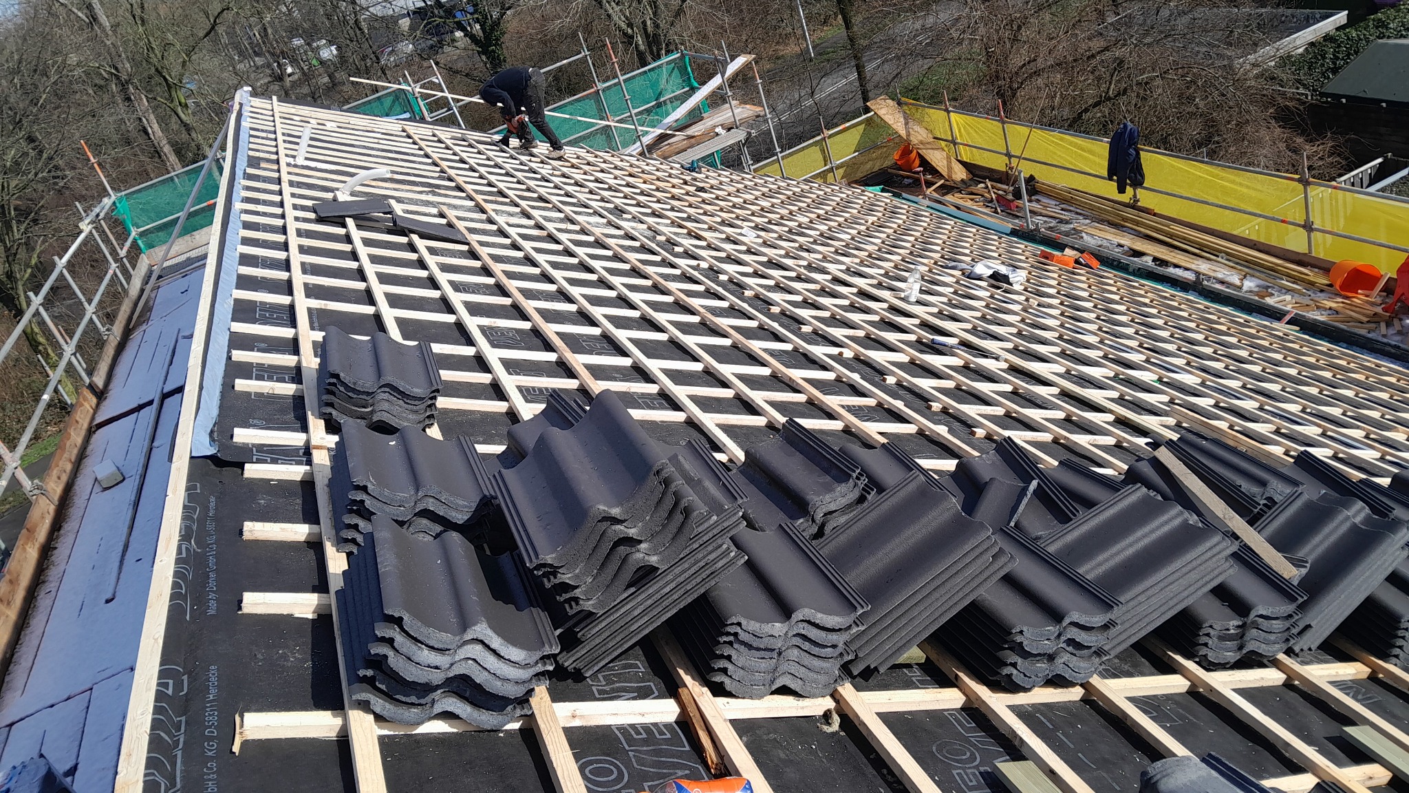 Układanie czarnych dachówek karpiówek na drewnianej konstrukcji dachu pokrytej membraną, w tle widoczny dekarz pracujący na dachu i rusztowania zabezpieczające.