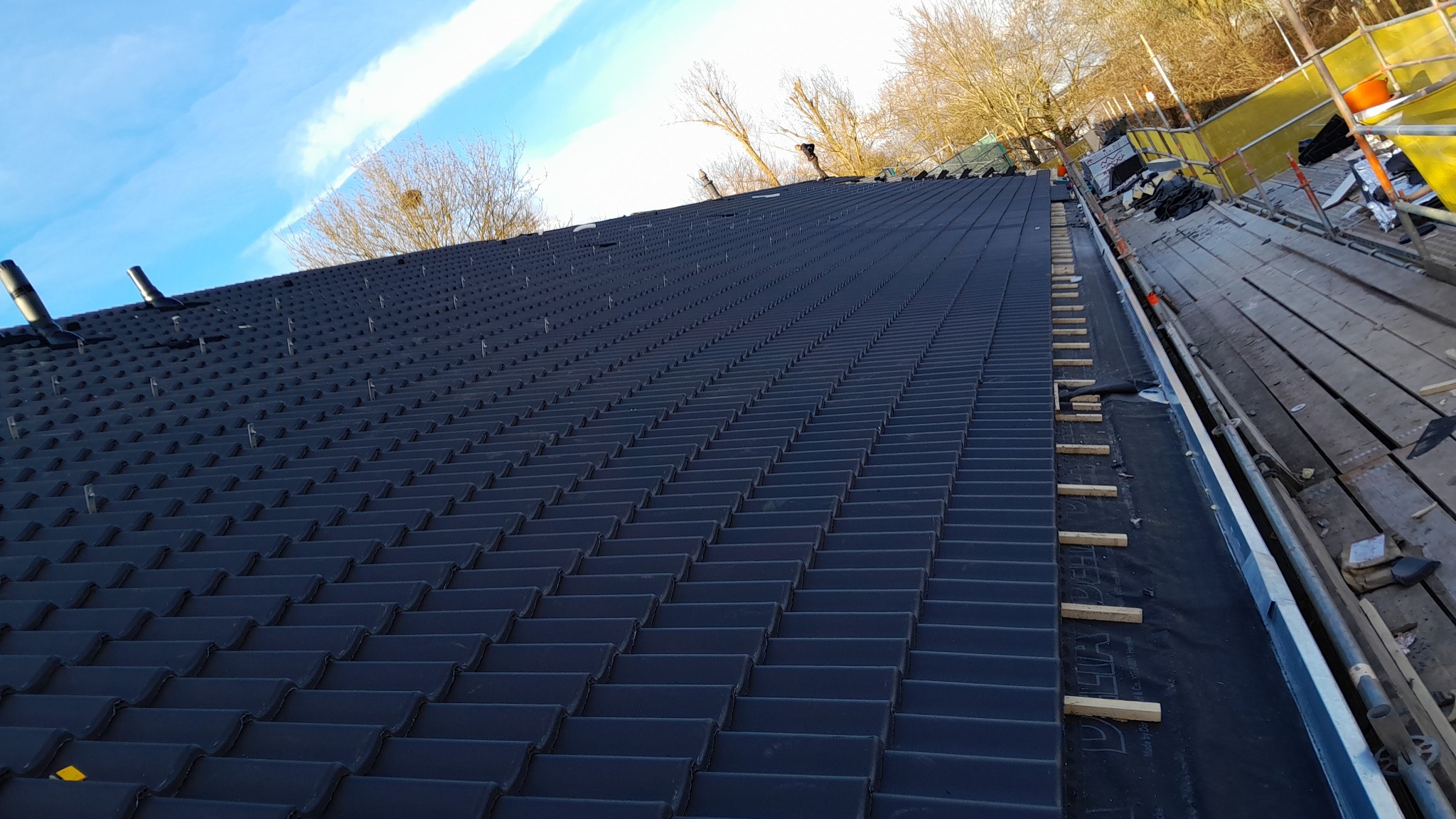 Nowy, czarny dach z dachówki ceramicznej w trakcie układania, widoczne elementy mocujące, rusztowanie i fragment nieba.