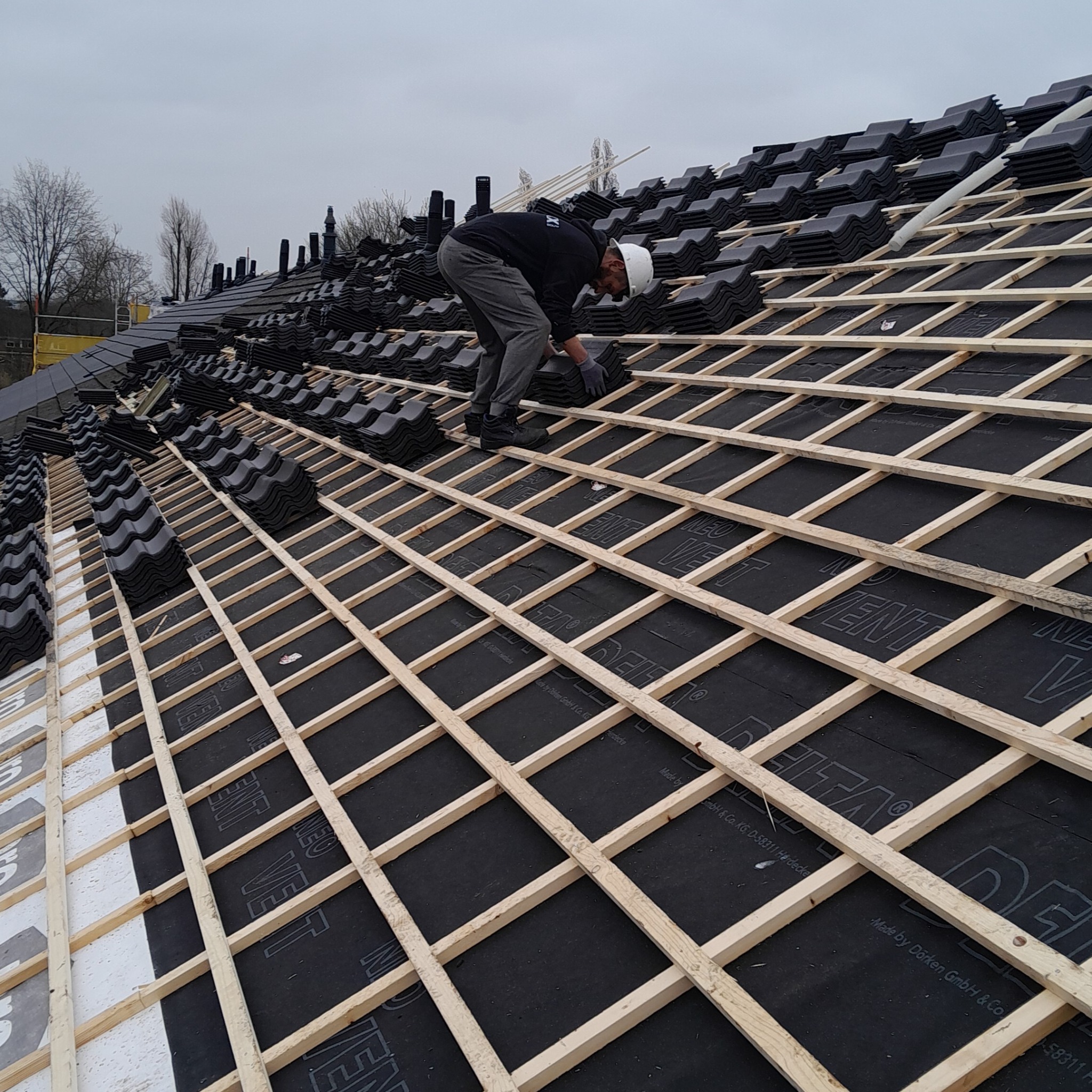 Pracownik w kasku układa ciemne dachówki na drewnianej konstrukcji dachu pokrytej czarną membraną z widocznym nadrukiem producenta.