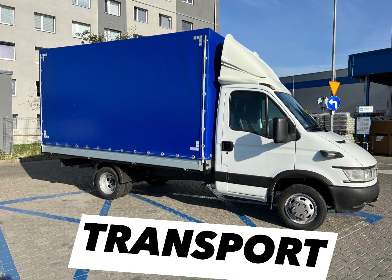 Biały samochód dostawczy z niebieską plandeką zaparkowany na parkingu z niebieskimi liniami, widoczny znak drogowy.