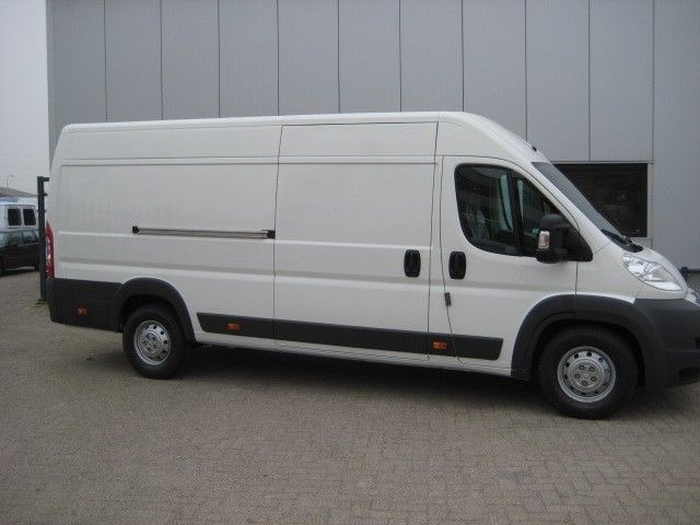 Biały, dostawczy bus marki Peugeot Boxer, zaparkowany na zewnątrz budynku o szarych ścianach, widziany z boku.