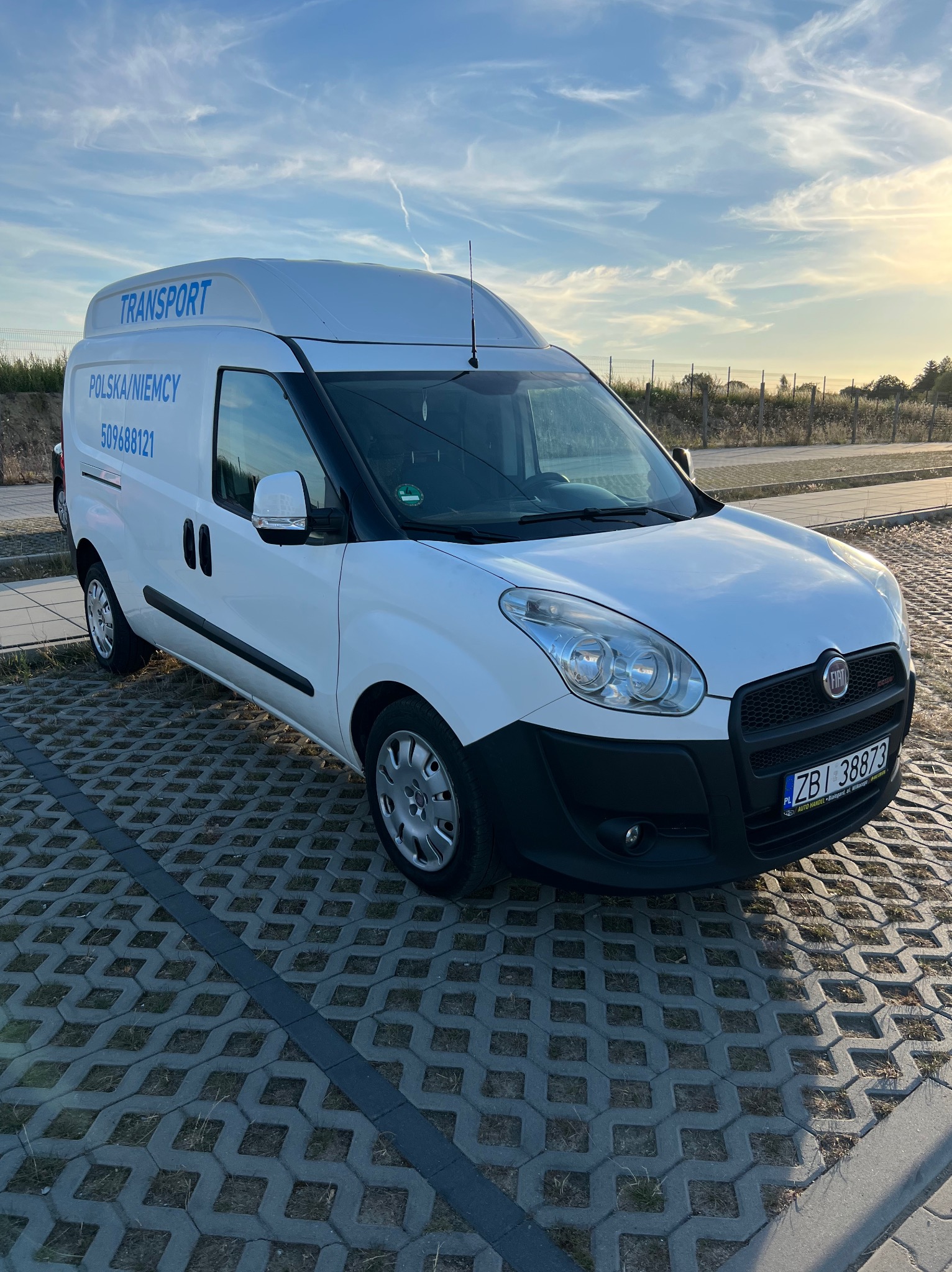 Biały Fiat Doblo z wysokim dachem i napisem 'TRANSPORT Polska/Niemcy 509688121' zaparkowany na ażurowym parkingu, widok z boku, w tle zachodzące słońce.