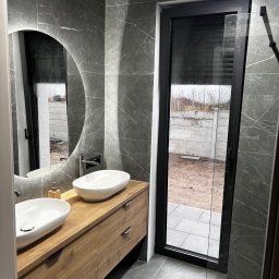 Sylwek Drętkiewicz „GLAZURNIK” - Nowoczesna łazienka z dwiema owalnymi umywalkami nablatowymi na drewnianej szafce, okrągłe lustro z podświetleniem LED i szare płytki imitujące marmur na ścianach i podłodze.